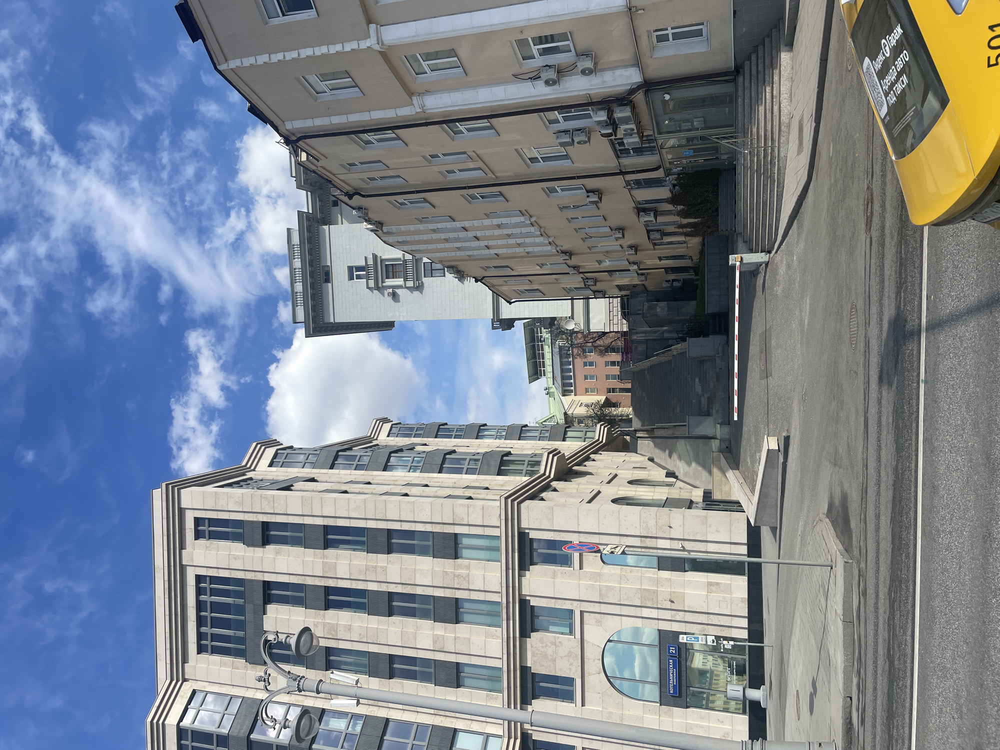 A street in Moscow, Russia, is framed by buildings on either side. The sky above is blue with scattered white clouds. The building on the left is a modern structure with a light-colored facade and rows of dark-framed windows. A street sign on the building reads "KOTELNICHESKAYA NAB. 21". A street lamp stands near the base of the building. On the right is an older, multi-story building with light-beige siding and many windows, some with air conditioning units. A yellow taxi is partly visible in the upper right corner. A set of concrete steps leads up from the street to a building entrance. A white security barrier is positioned across the street.