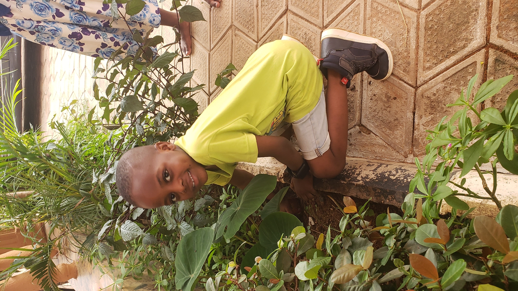 A young boy in a lime green shirt, gray shorts, and dark shoes is playing in a garden in what appears to be Narayi, Nigeria. He is bent over, looking at the camera with a smile. Various plants and greenery surround him. A wall of hexagonal concrete blocks forms the background on one side, and the lower portion of a woman's dress with blue floral patterns is visible on the other. The setting appears to be outdoors, possibly a yard or garden adjacent to a building.