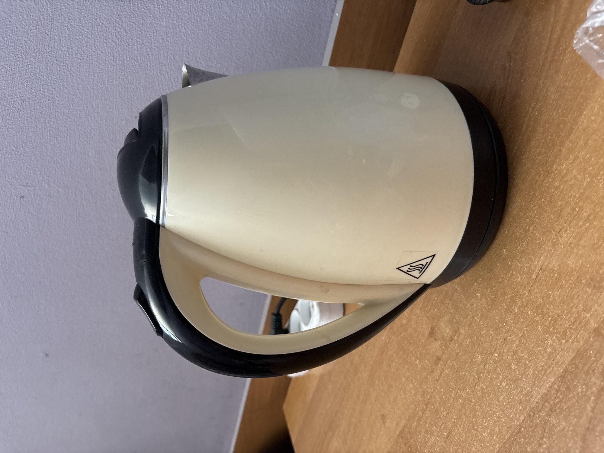 The image shows a close-up, angled shot of a cream-colored electric kettle with a black handle and trim. The kettle is positioned on a wooden surface, possibly a table or counter. To the left of the kettle is a light purple, textured wall. The lighting suggests it could be daytime, though no windows or external light sources are visible. The scene is a still life, with the kettle being the sole focus. A small triangular warning symbol with wavy lines indicating heat is visible on the side of the kettle. No people are present in the image, and no specific activity is taking place. The overall impression is one of a domestic or office setting, with a focus on a common household appliance.