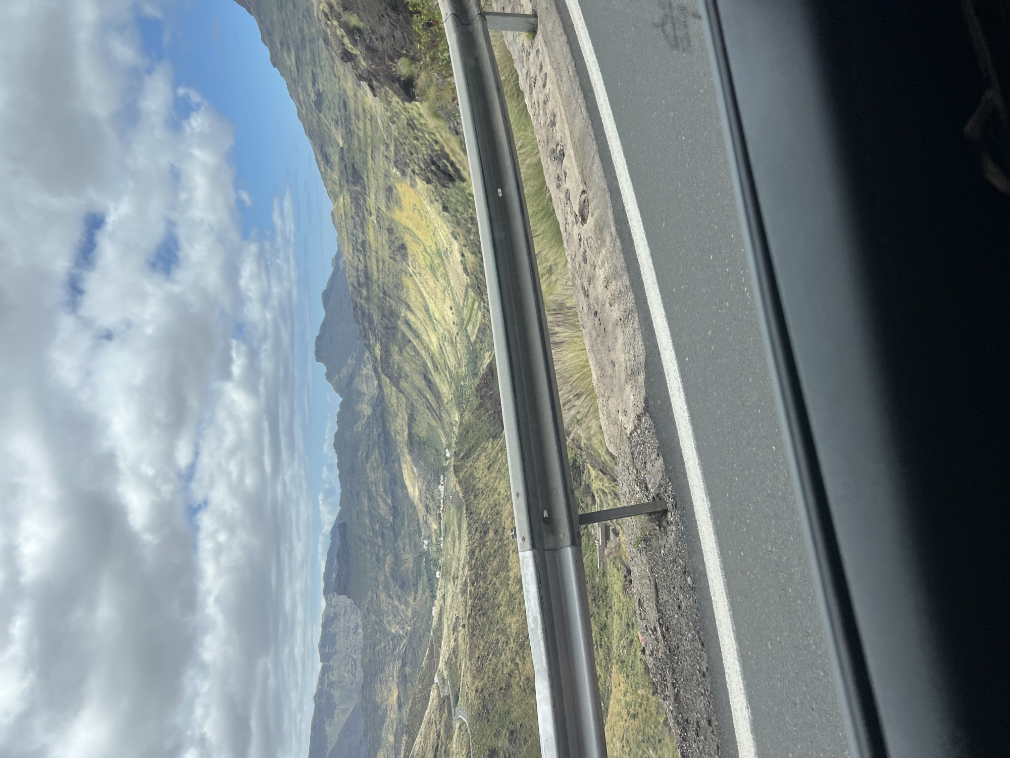 The image captures a view from a vehicle moving along a road in Mogán, Spain. The landscape is mountainous with varied vegetation. The sky is partly cloudy with blue patches visible. A guardrail runs parallel to the road. The vehicle's interior is visible on the right side of the frame, suggesting the image was taken from inside the car.