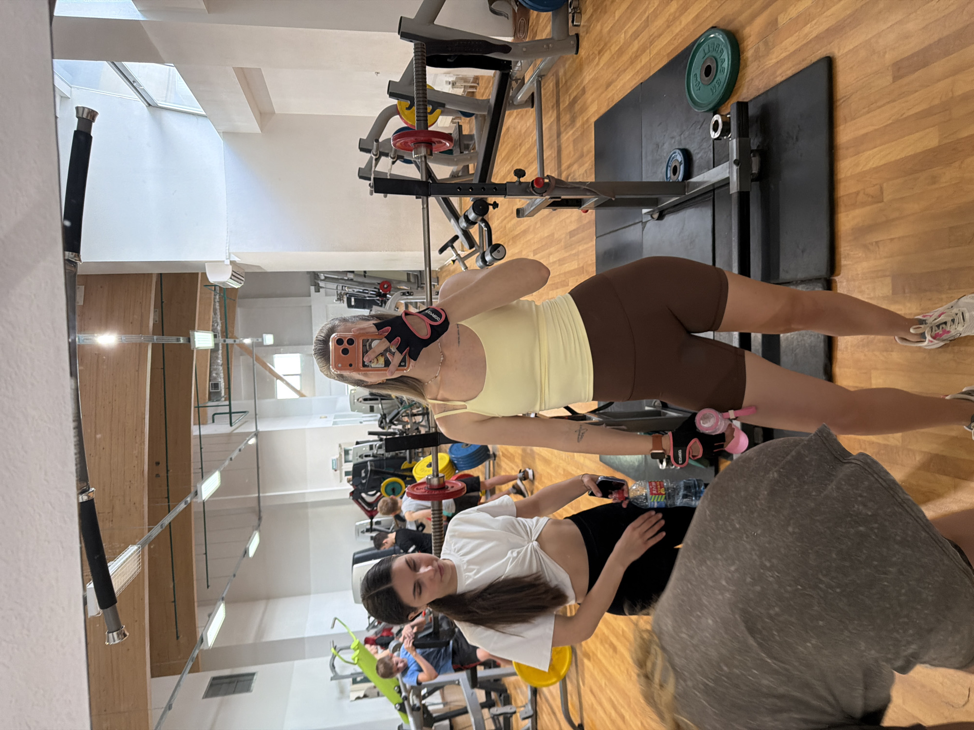 In a brightly lit gym with wooden floors, two women are visible. The woman on the right, in the foreground, is taking a mirror selfie. She is wearing a light yellow tank top and brown shorts. She has blonde hair and is wearing black and pink workout gloves. She is holding a cell phone in her right hand. To her left, another woman with dark hair is looking down and holding a water bottle. She is wearing a white knotted crop top and black shorts. In the background, several men and gym equipment, including weight racks and barbells, are visible. The gym appears to be located in Podkletnoe, Russia.