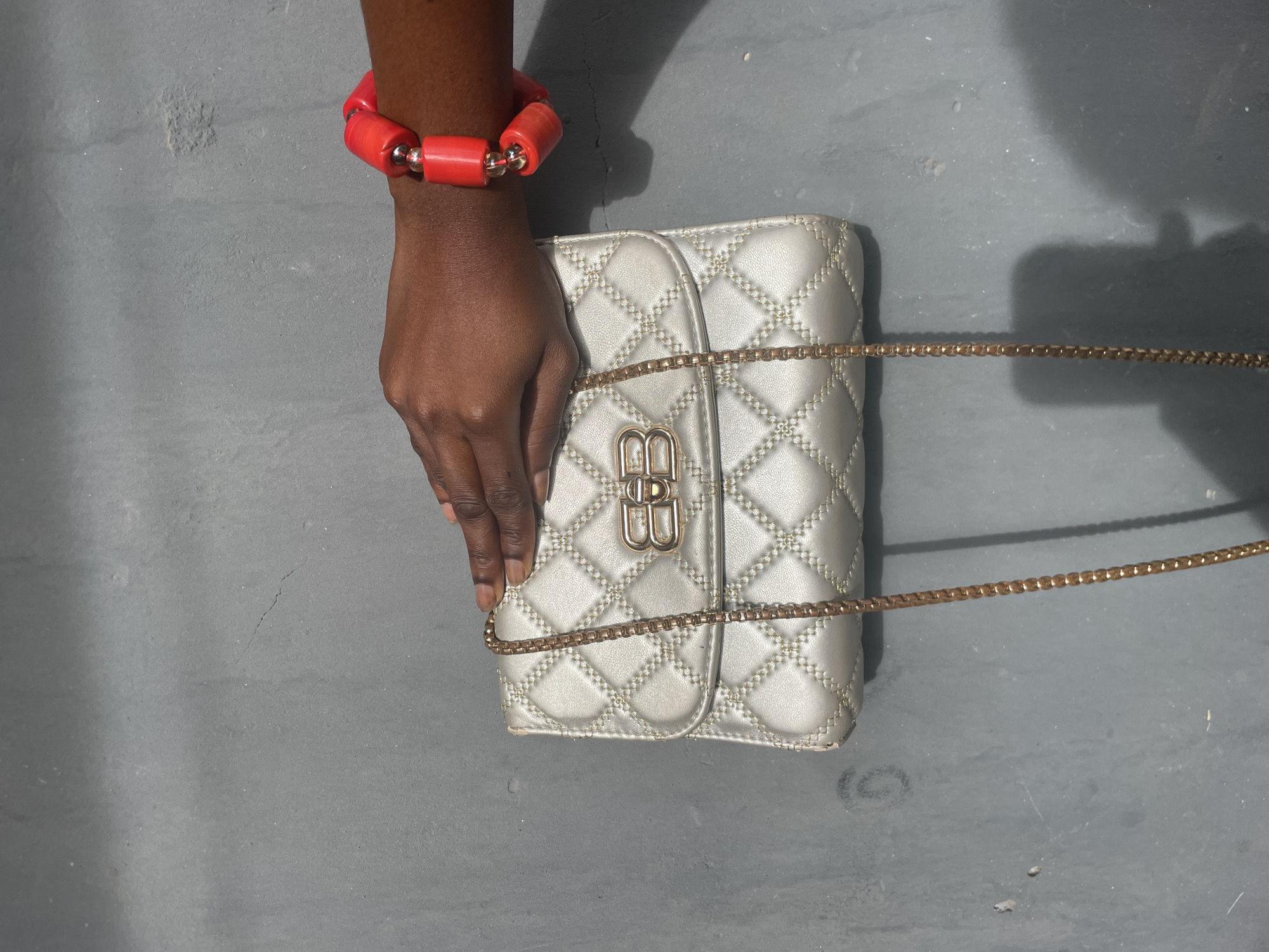 A close-up, high-angle shot captures a dark-skinned hand adorned with a vibrant red and silver beaded bracelet, holding a small, quilted handbag against a textured gray surface. The scene is set outdoors in daylight, likely in Badagry, Nigeria, given the strong shadows cast by the hand and an unseen object, suggesting bright sunlight.
The hand, with neatly manicured nails, gently grasps the light metallic, possibly silver or champagne gold, handbag. The bag features a diamond-shaped quilted pattern with visible stitching and is accented with a delicate gold-colored chain strap. A prominent gold-toned clasp on the front of the bag displays two intertwined 'B's, characteristic of the Balenciaga logo. The red bracelet, composed of cylindrical coral-like beads separated by smaller spherical silver beads, adds a striking pop of color against the neutral tones of the bag and background. The gray surface beneath is somewhat uneven and shows minor cracks and imperfections, consistent with concrete or a similar outdoor material.