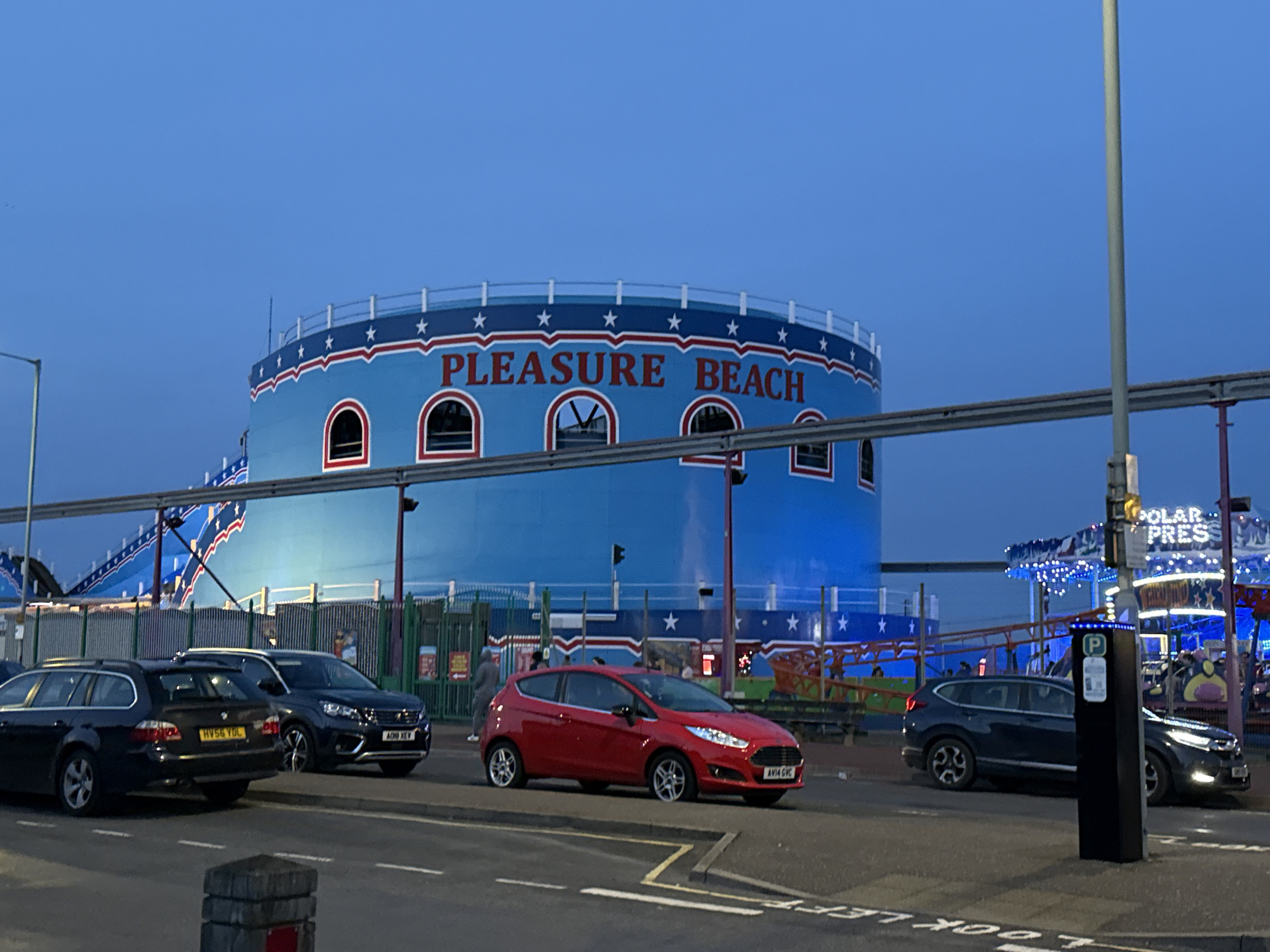 The image depicts a street scene in Gorleston-on-Sea, United Kingdom, with the prominent "Pleasure Beach" structure in the background. The building is cylindrical, painted blue with red and white accents including stars and the words "PLEASURE BEACH" in red. In the foreground, several cars are parked along the roadside. The vehicles include a dark-colored estate car (HV56 YOL), a dark SUV (AOM XEV), a red hatchback (AV14 GVC), and a dark SUV. Street lights and overhead structures are visible. A parking meter/pay station is to the right. The overall lighting suggests it is dusk. A portion of a fairground ride, marked "POLAR PRESS," is visible in the background to the right. The road has "LOOK LEFT" marked in white paint.