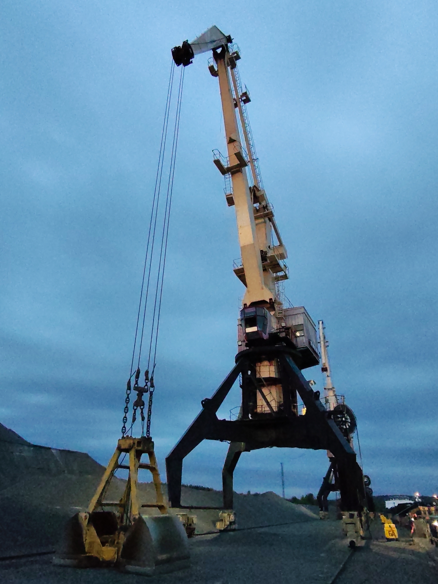 The image depicts a large harbor crane against a cloudy sky, likely at dusk or dawn due to the subdued lighting. The crane has a distinctive yellow clamshell bucket suspended by cables and chains. The crane's primary structure is a beige color, while its support legs are black. The location appears to be an industrial area in or near the city of Shalsky, Russia, considering the presence of what seems to be piles of aggregate and some other industrial infrastructure in the background. There is no indication of people in the immediate foreground, but there are lights in the distant background, implying some activity.