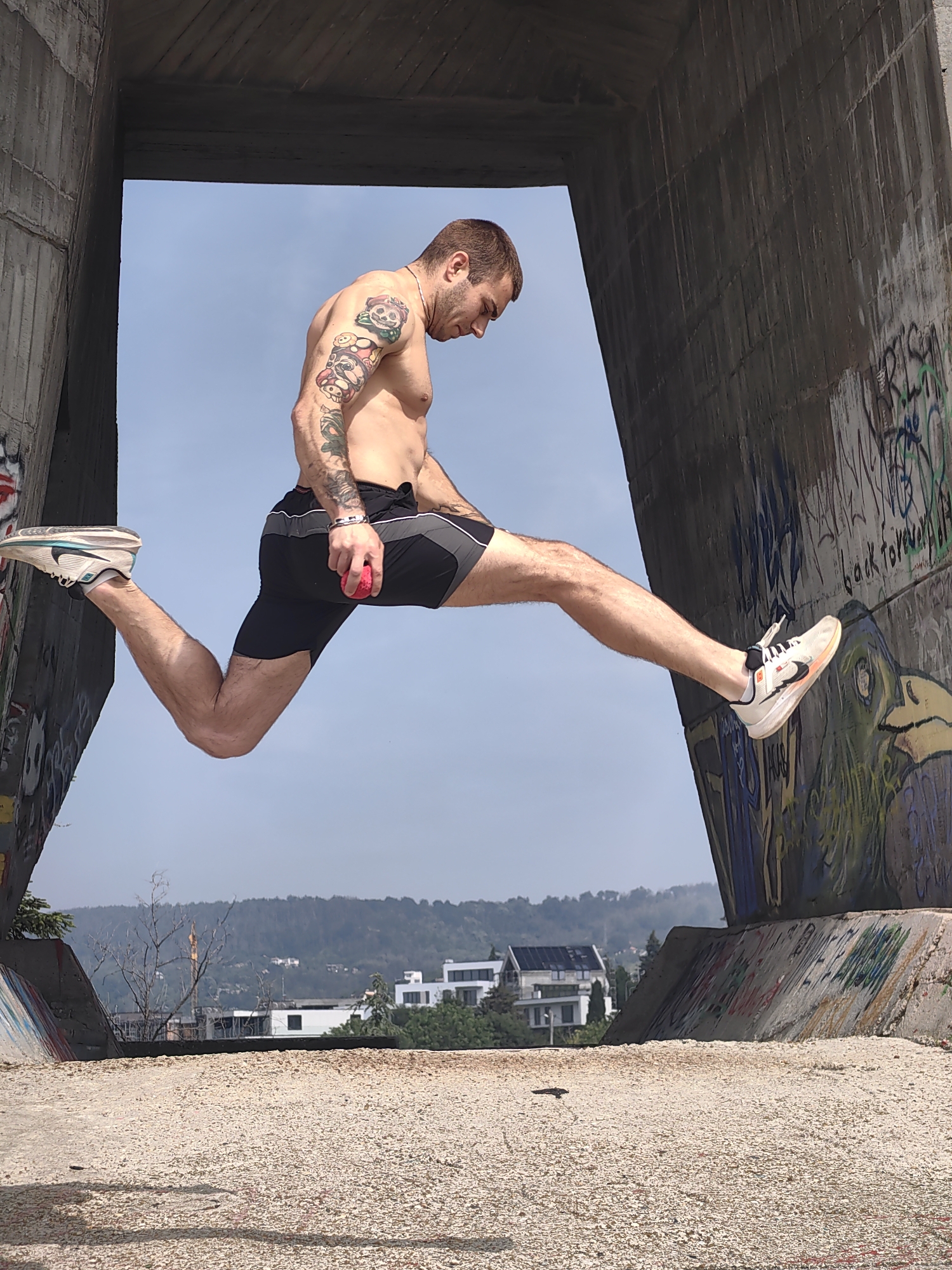 The image displays a male subject suspended in mid-air within a large, weathered concrete archway. The subject is shirtless, revealing a muscular physique, short brown hair, and stubble. He possesses extensive, colorful tattoo sleeves on both arms, depicting various cartoon-like characters and designs. He is dressed in black athletic shorts with grey side panels worn over black compression shorts, and white athletic shoes with black, orange, and light blue accents. A silver chain necklace and a silver bracelet on his left wrist are visible. His body is oriented horizontally; the right leg is extended forward and slightly downward, while the left leg is sharply bent backward, foot angled upward. His head is tilted downwards, and his right hand grasps a small, spherical, textured red object.
The concrete structure forming the archway is aged and textured, exhibiting vertical streaks and patches of discoloration. Both the interior and exterior surfaces of the concrete walls are covered in extensive, multicolored graffiti, which includes various tags, stylized lettering, and larger artistic renditions, such as a green, bird-like creature on the lower right wall. The ground beneath the subject and extending through the archway consists of light-colored gravel or small pebbles.
Beyond the archway, a clear, light blue sky dominates the upper background. Below the sky, a panoramic view of distant, tree-covered hills unfolds. Several modern white buildings, some featuring dark roofs and solar panels, are nestled among the trees on these hills. A few bare trees are visible in the middle ground, situated closer to the concrete structure. The scene is brightly lit by natural daylight. This urban-natural landscape, characterized by hills and contemporary architecture, is consistent with descriptions of Varna, Bulgaria.