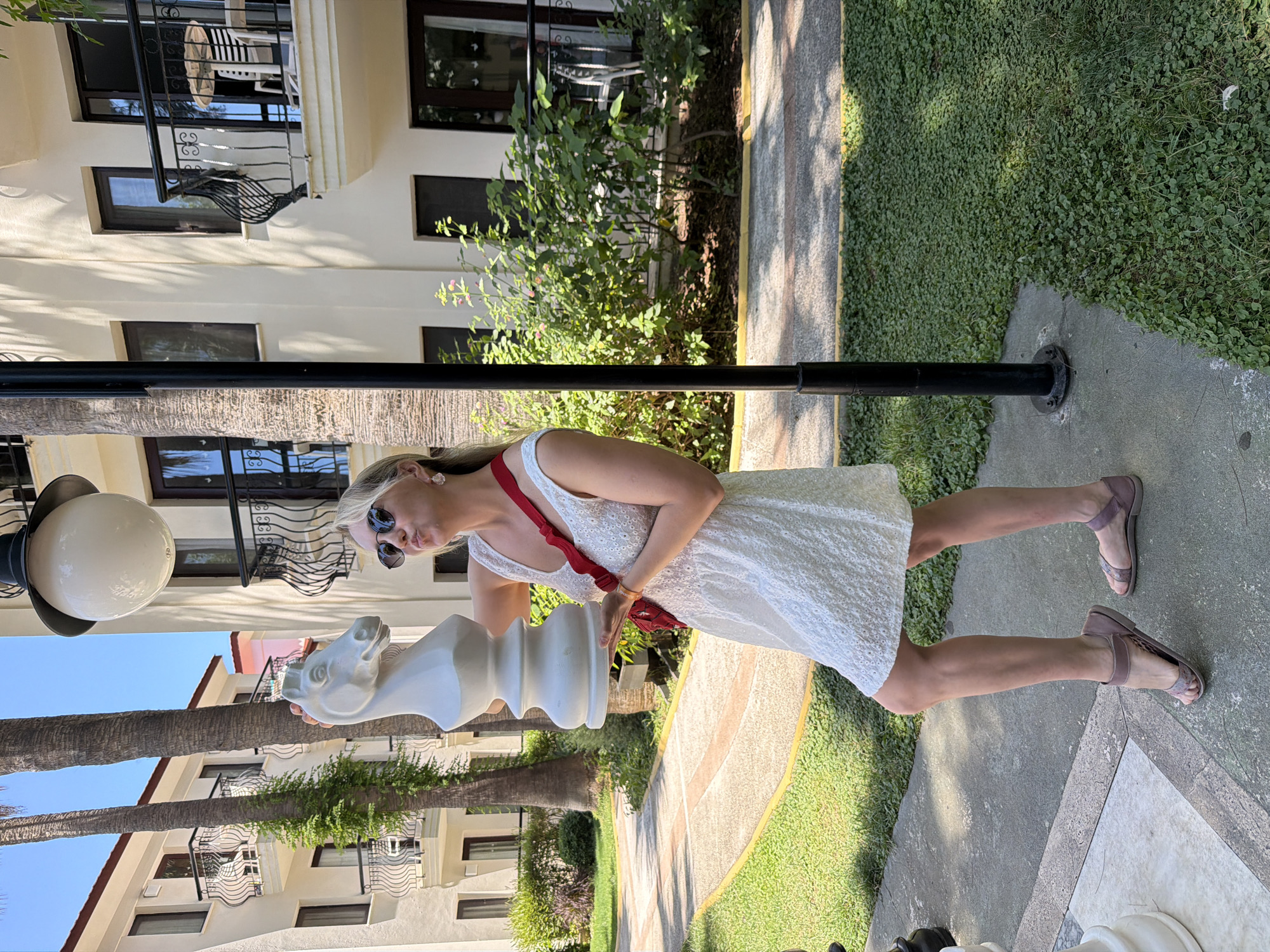 A woman stands on a paved path next to a lawn in Fethiye, Turkey. She wears a white dress and sunglasses and holds a large, white chess knight piece. A black pole with a round light fixture is to her left. Buildings with windows and wrought-iron balconies are in the background, along with trees and foliage.