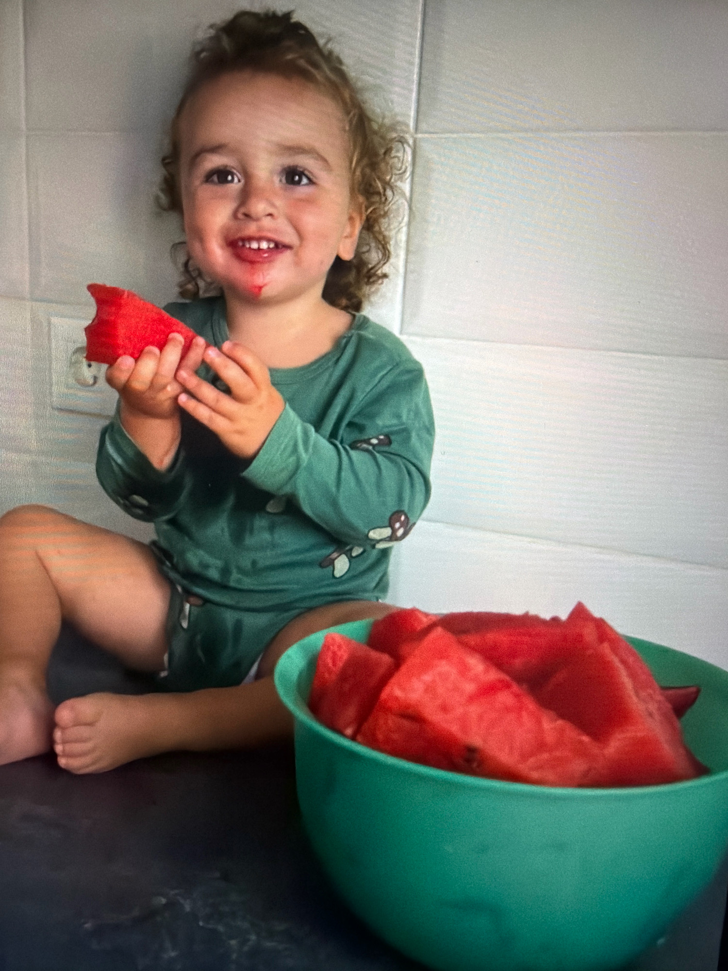 A young child with curly brown hair and a happy expression sits in front of a bowl of watermelon slices. The child is wearing a green long-sleeved shirt and appears to have watermelon juice around their mouth. They are holding a slice of watermelon in their hands. A large green bowl filled with watermelon pieces is positioned to the child's right. The background consists of a white tiled wall. The setting appears to be a kitchen. There is no information about the city of Vniissok, Russia present in the image.