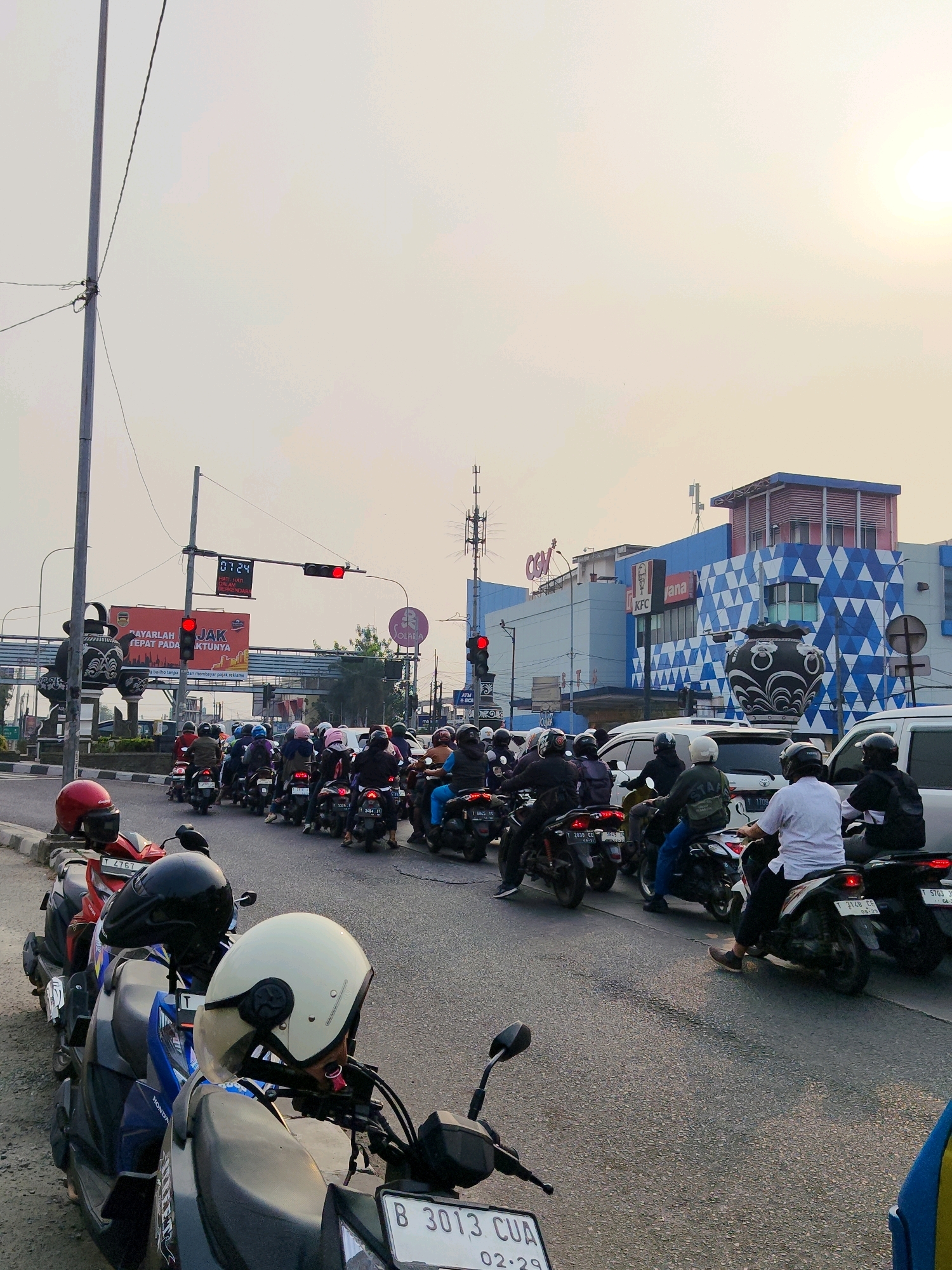 The image depicts a street scene in Purwakarta, Indonesia, at an intersection. A line of motorcycles is stopped at a red traffic light, with several cars also present in traffic. The sky is overcast with a bright sun in the upper right corner. A tall pole with electrical wires is on the left side of the frame. A traffic signal displays a red light. A digital timer above the traffic light shows "07:24". A large billboard with text in Indonesian and an image of a decorative vase is visible behind the traffic light. Further down the street, a building with a blue and white geometric pattern is prominent, identified by signs for "CGV" and "KFC". People are seated on the motorcycles, mostly wearing helmets. The foreground shows the handlebars and seats of several motorcycles, with helmets placed on some. The road surface is asphalt.