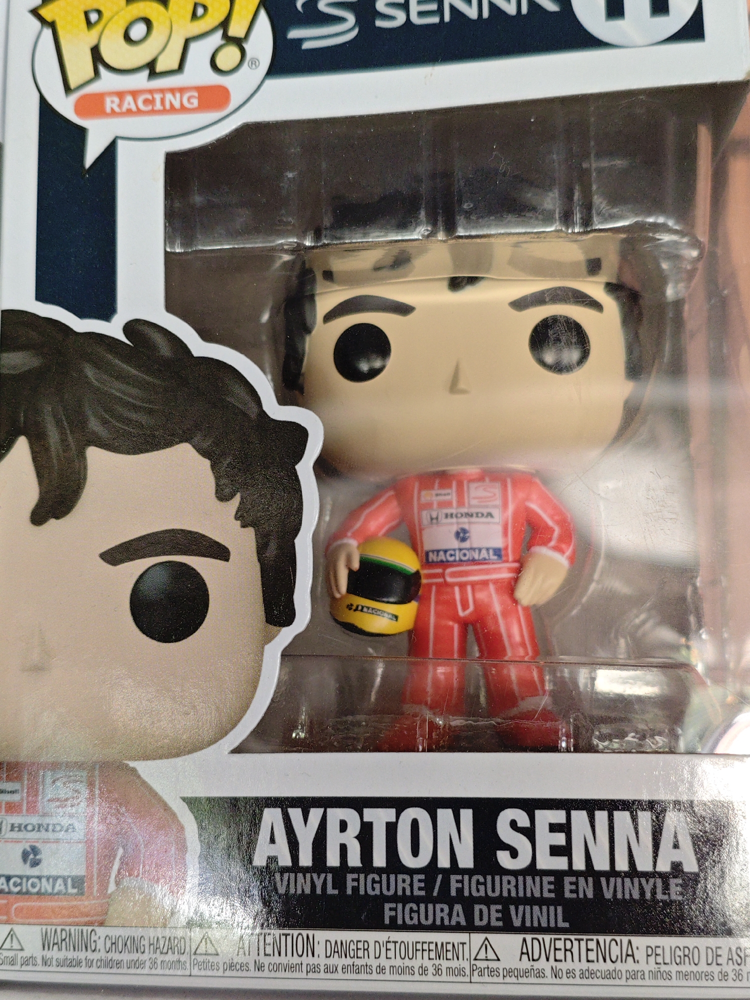 The image shows a Funko Pop! racing figure of Ayrton Senna, a renowned Formula 1 driver. The figure is encased in its original plastic packaging, which has some minor scuffs and scratches. The packaging itself features the "Pop!" logo and the word "RACING" in an orange speech bubble. Below the figure, the packaging displays "AYRTON SENNA" in large white letters, with smaller text indicating "VINYL FIGURE / FIGURINE EN VINYLE / FIGURA DE VINIL".
The figure itself depicts Ayrton Senna in his iconic red racing suit with white stripes. He is holding a yellow and black racing helmet. The figure's face is characteristic of the Funko Pop! style, with large black circular eyes and a simple nose and mouth.
The background of the image is blurry but appears to be an indoor setting, possibly a room or a shelf. There is no visible indication of the location being in the city of Kyiv, Ukraine. The lighting suggests an artificial light source.