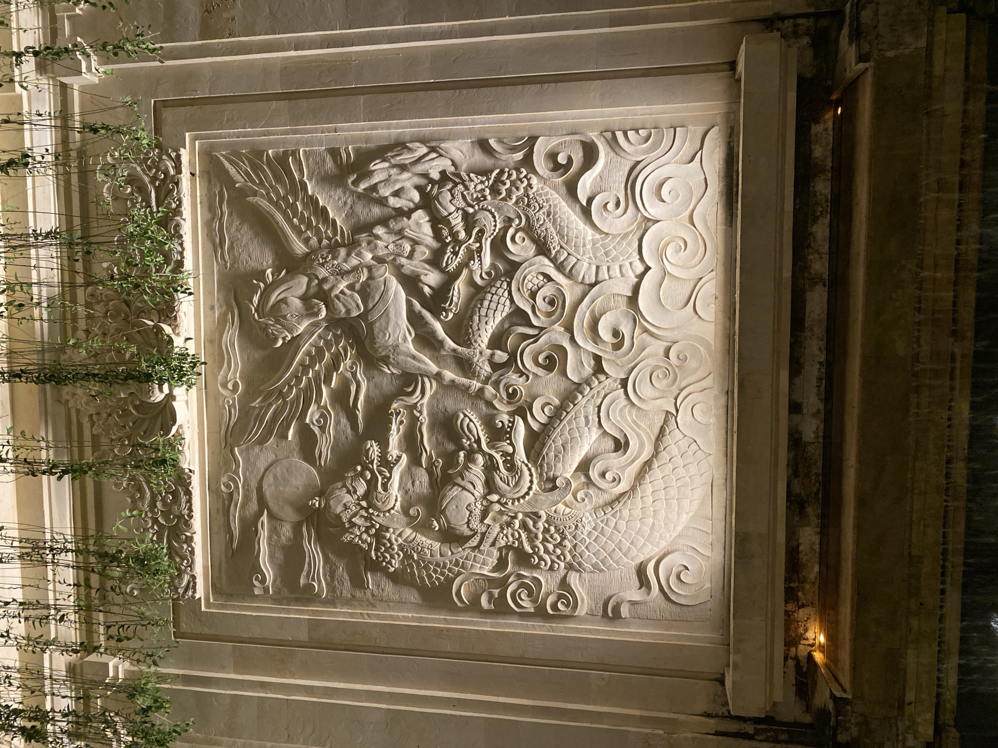 The image shows a large, intricately carved stone relief illuminated by artificial lights. The relief depicts a mythical scene with a winged horse, possibly Pegasus, at the center, flying above two dragons. The dragons are intertwined and appear to be in combat or motion, with swirling clouds and waves rendered around them. The artwork is mounted on a textured wall, with decorative elements and cascading green vines hanging from above. Below the relief, a stone ledge is visible, with a faint glow from a hidden light source. Further down, a dark area suggests a water feature or pool, with streaks of light indicating falling water. The overall impression is of an outdoor decorative element, possibly part of a building's facade or a garden feature, situated in Kuta Selatan, Indonesia.