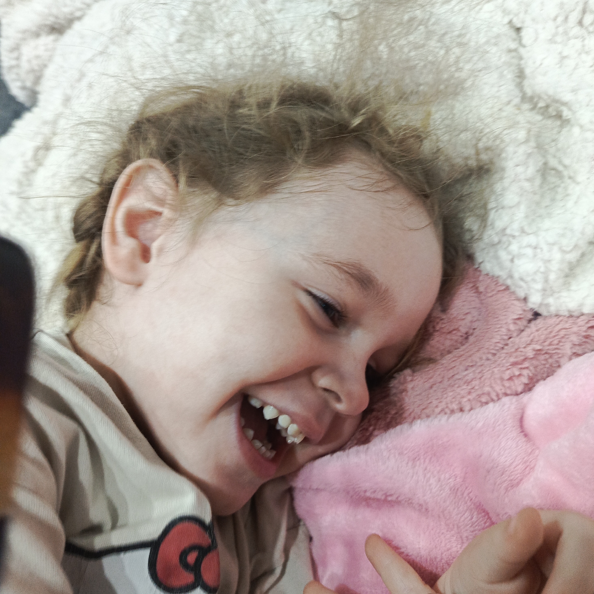 A young child lies on a white, fluffy blanket and a pink, furry pillow, smiling broadly with visible teeth. The child has light brown hair braided on the side. The child is wearing a light-colored shirt with a red and black design on it. The location is Марковский, Russia.