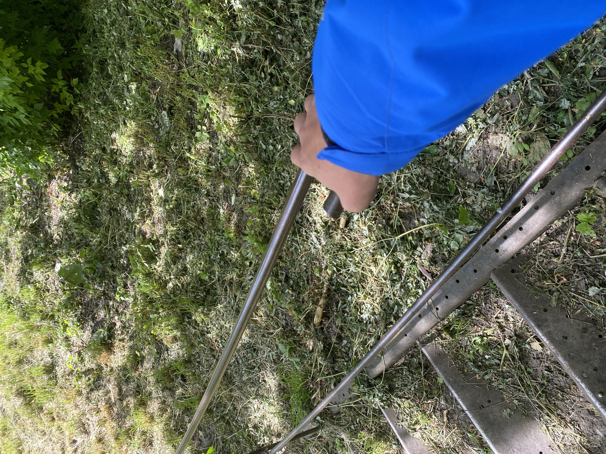 A hand wearing a bright blue sleeve is grasping a silver metal railing, next to a metal staircase on overgrown grassy ground, located in an unspecified part of Ustyvytsia, Ukraine. The scene appears to be outdoors.