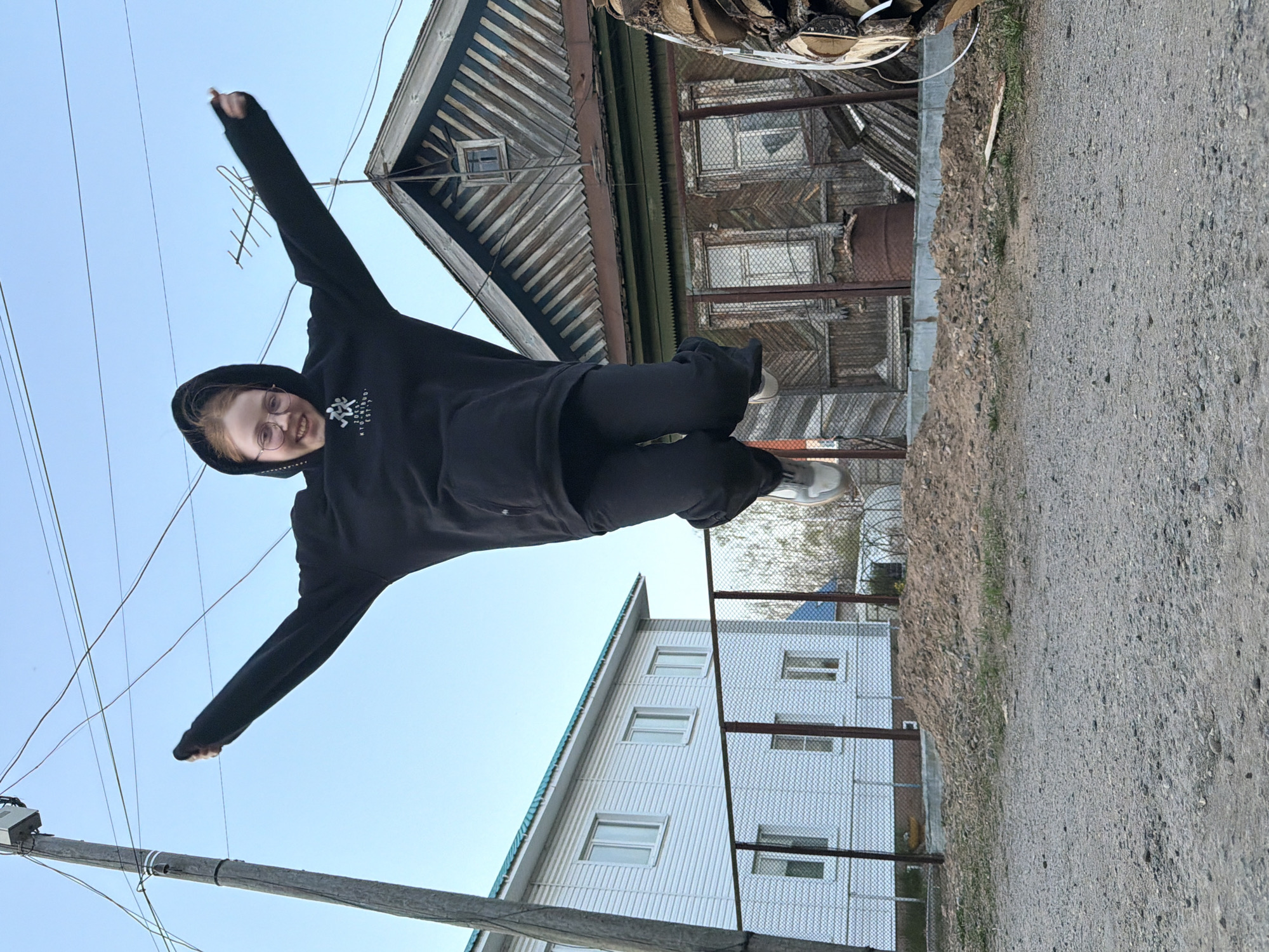 A person is mid-air with outstretched arms, appearing to jump or float. The person is wearing black clothing. The background includes a gravel path, two buildings (one old with wood paneling and one newer with white siding), a fence, power lines and a pole. This appears to be near Chaikovsky, Perm Krai, Russia.