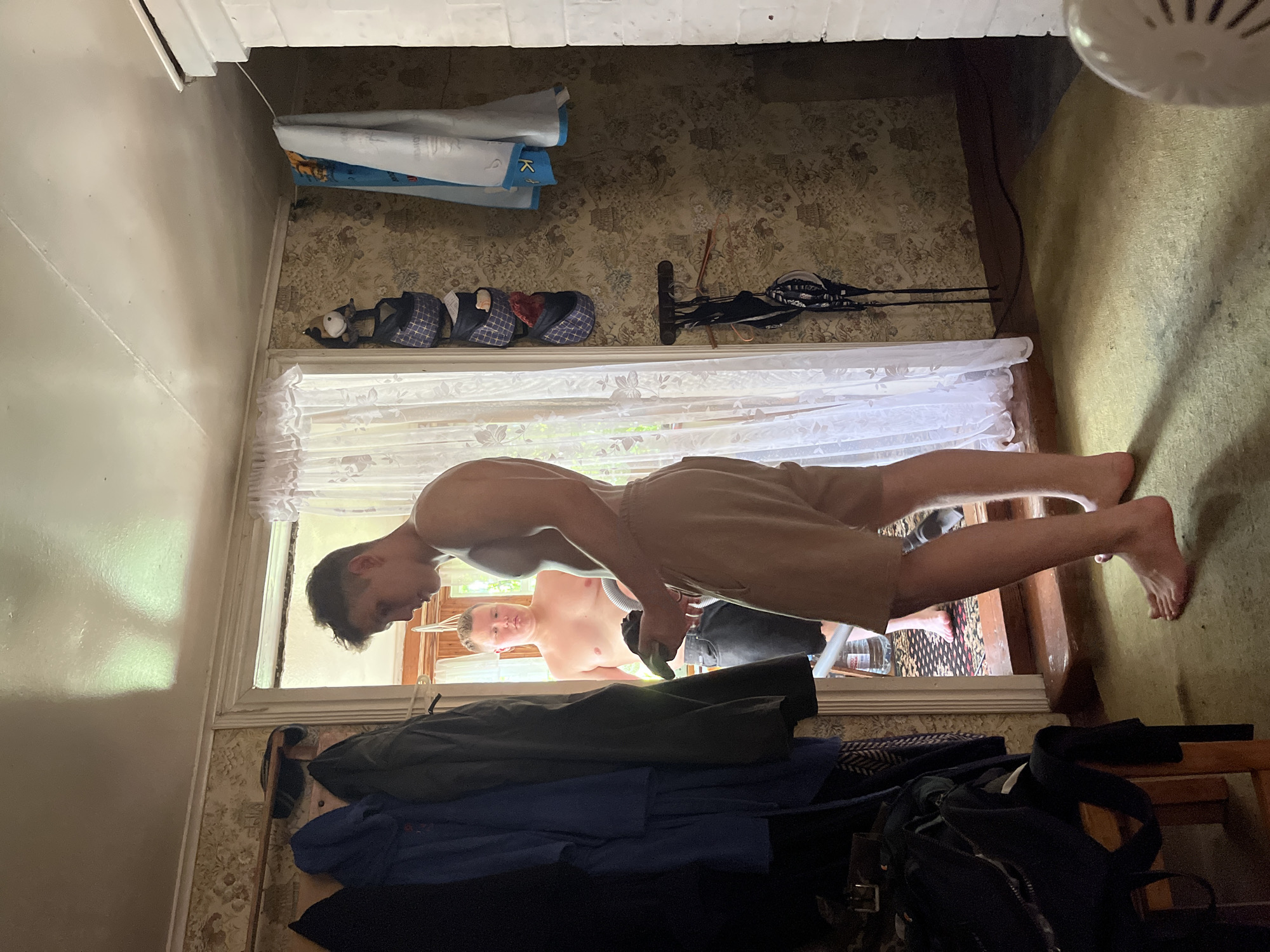 A young man, appearing to be in his late teens or early twenties, stands in the foreground facing away from the camera, looking towards an open doorway. He is shirtless and wearing light-colored shorts. In his hands, he holds a dark object and a green item, possibly a piece of clothing or a towel. Through the doorway, another young man, also shirtless, is visible, wearing dark shorts. He stands near a bottle of water on the floor. The room appears to be a rustic interior, possibly a rural house. Coats are hanging on a rack to the left. The walls are covered in patterned wallpaper. A white curtain is visible in the doorway. The floor is carpeted in a light green color. The lighting suggests it is daytime, with sunlight streaming through the doorway. There is a white structure on the right side of the frame, possibly a stove or fireplace. The scene is candid and informal. The location is the city of Sad. Uchastki, Russia.