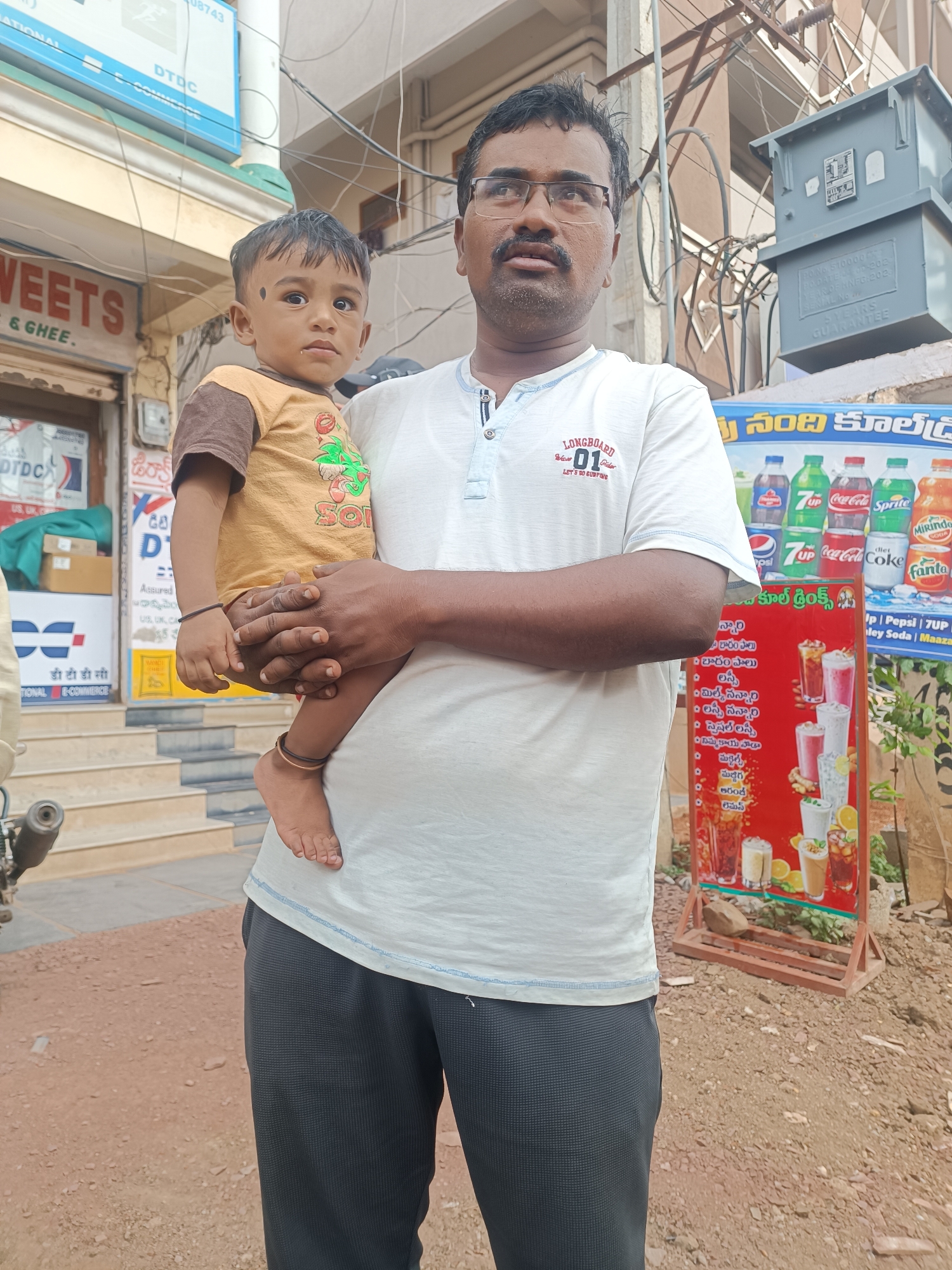 The image captures a man in Bethamcherla, India, holding a young child in his arms. The man, wearing glasses and a white t-shirt, stands on a dusty street. The child, dressed in a yellow and brown shirt, has a dark mark on its forehead. Behind them, a shop displays various beverage brands, including Coca-Cola and Sprite. An electrical transformer is visible on a pole in the background. The scene appears to be a street-side view of a commercial area in Bethamcherla.