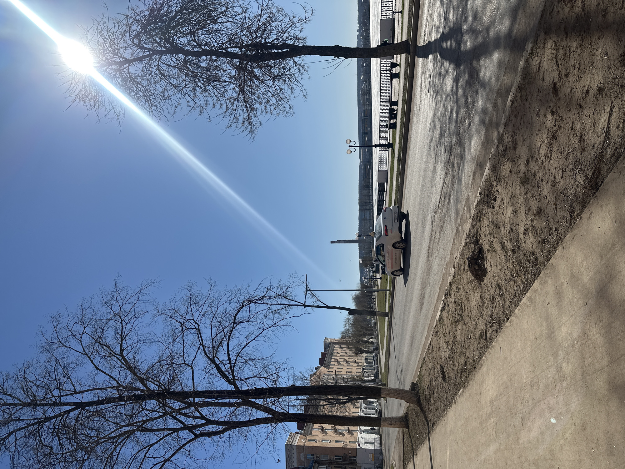 The image shows a street scene in what is likely Votkinsk, Russia, based on contextual cues like architecture and vehicle license plates. The view is along a roadside, with a sidewalk in the foreground transitioning to a dirt median. A roadway is present where a white car is driving. Beyond the road, there is a fence, what appears to be a pedestrian walkway along a body of water, and an industrial complex. The sky is blue with the sun prominently visible, creating a bright glare. Two bare trees are positioned on each side of the image.