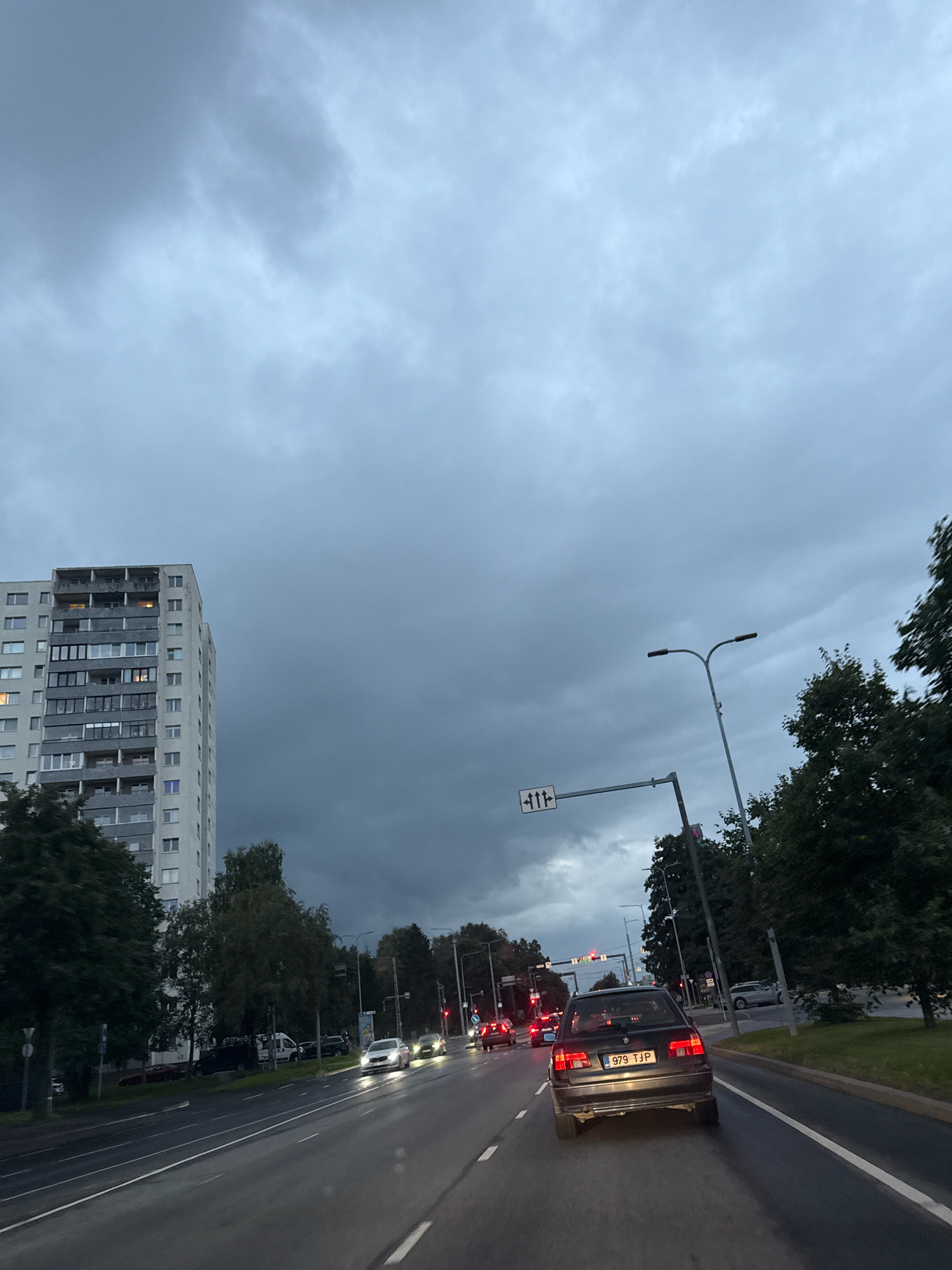 A car with license plate 979 TJP is driving on a wet, multi-lane road in Tallinn, Estonia, under a cloudy, overcast sky. The car is a dark-colored sedan, facing away from the camera. In the background, there is a tall, grey apartment building with many windows and balconies on the left side of the frame. Trees line the side of the road, and streetlights are visible. Traffic signals are present. Other vehicles are driving in the same direction, and some are approaching from the opposite direction. The lighting suggests it is either early morning or late evening, or a very dark, cloudy day.
