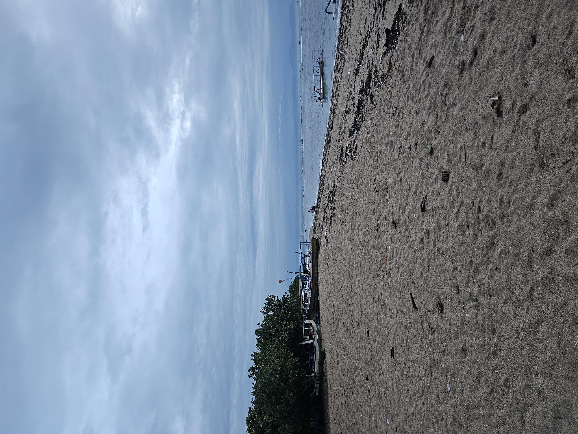 The image shows a beach in Denpasar, Indonesia, with a strip of sandy beach taking up much of the right side of the frame. The sand is a brownish-grey color. A line of debris is visible along the upper edge of the beach. The sky is a light blue with scattered clouds. The ocean appears as a light blue expanse leading to the horizon. In the middle ground, there are boats parked along the shoreline near some trees, one of which is marked with a red flag.
