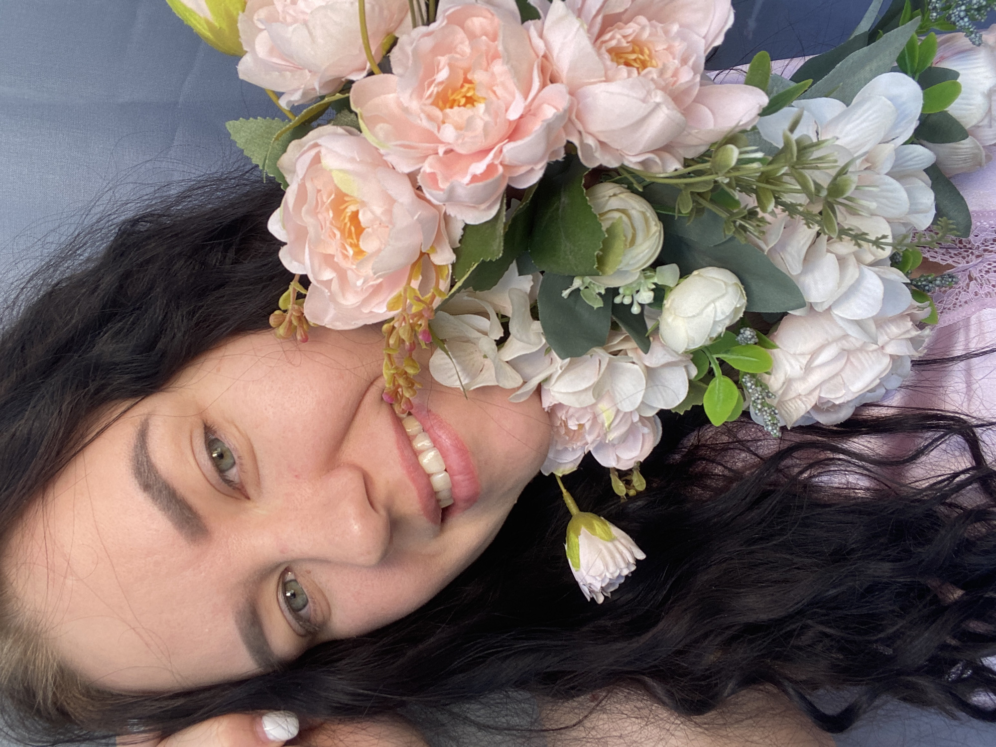 A woman with dark hair and fair skin lies on a gray surface, adorned with a crown of pink and white flowers with green leaves. She has light green eyes and is smiling, revealing her teeth. Her hair is long and wavy, with a hint of white on one section near her face. The location is not discernible from the image.
