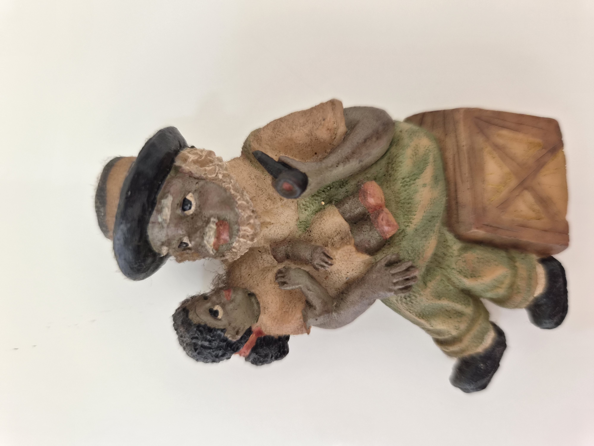 This is a photograph of a ceramic figurine of an elderly Black man holding a young Black child. The man is wearing a hat, a tan shirt, and green pants. He is sitting on a wooden crate and holding a pipe in his right hand. The child is wearing a tan dress and is being held in the man's left arm. The figurine is placed against a plain white background. The image does not depict any location in Maltepe, Turkey.
