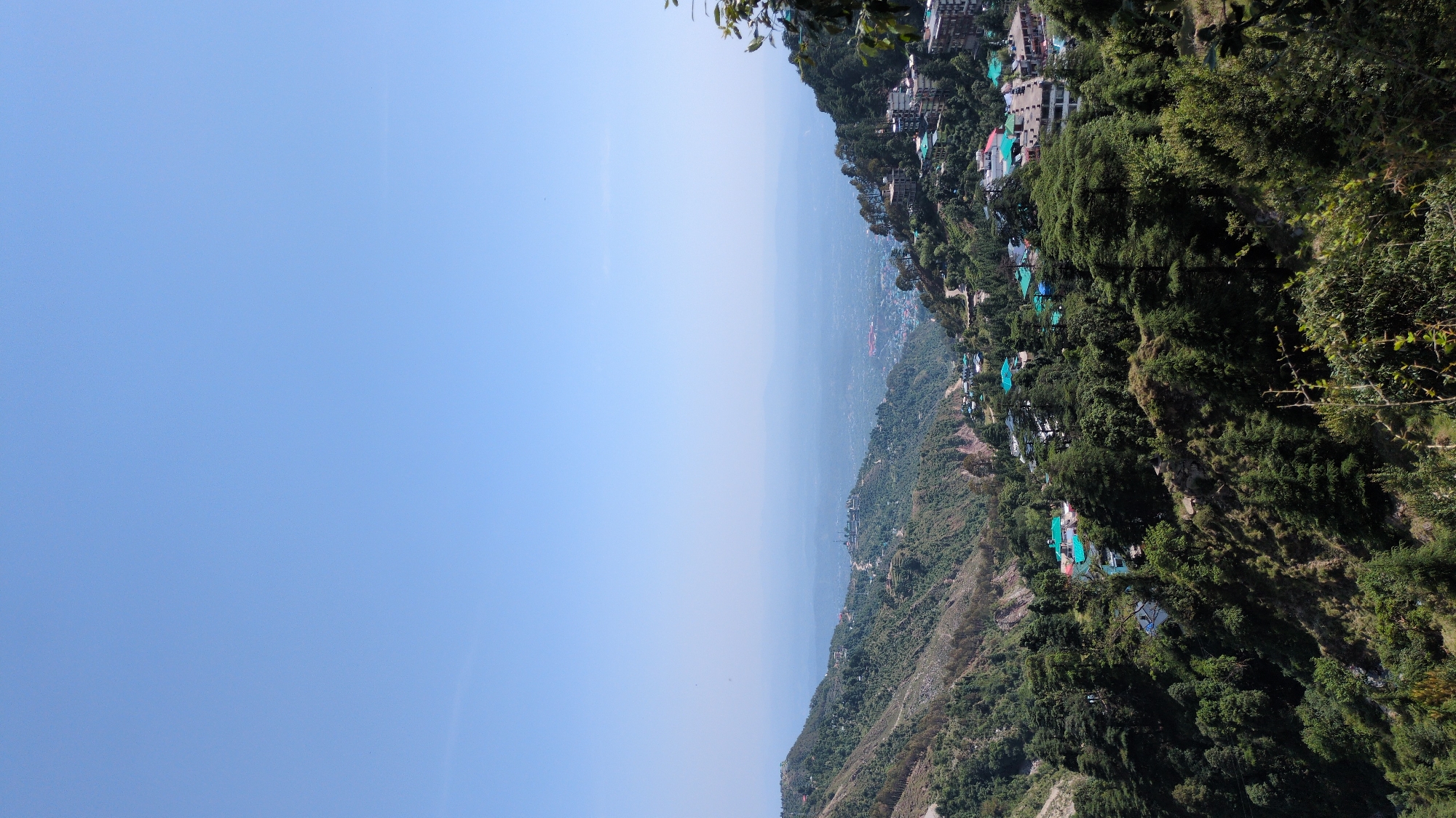 The image presents an elevated view of a hilly landscape, possibly in or near Bhagsu, India. The sky, predominantly clear blue, occupies the left portion of the frame. The right side is dominated by a hillside covered in dense vegetation and scattered buildings, some with distinctive turquoise roofs. A distant view of a valley or plain is visible between the hillside and the sky. The shot seems to have been taken during a clear, bright day.