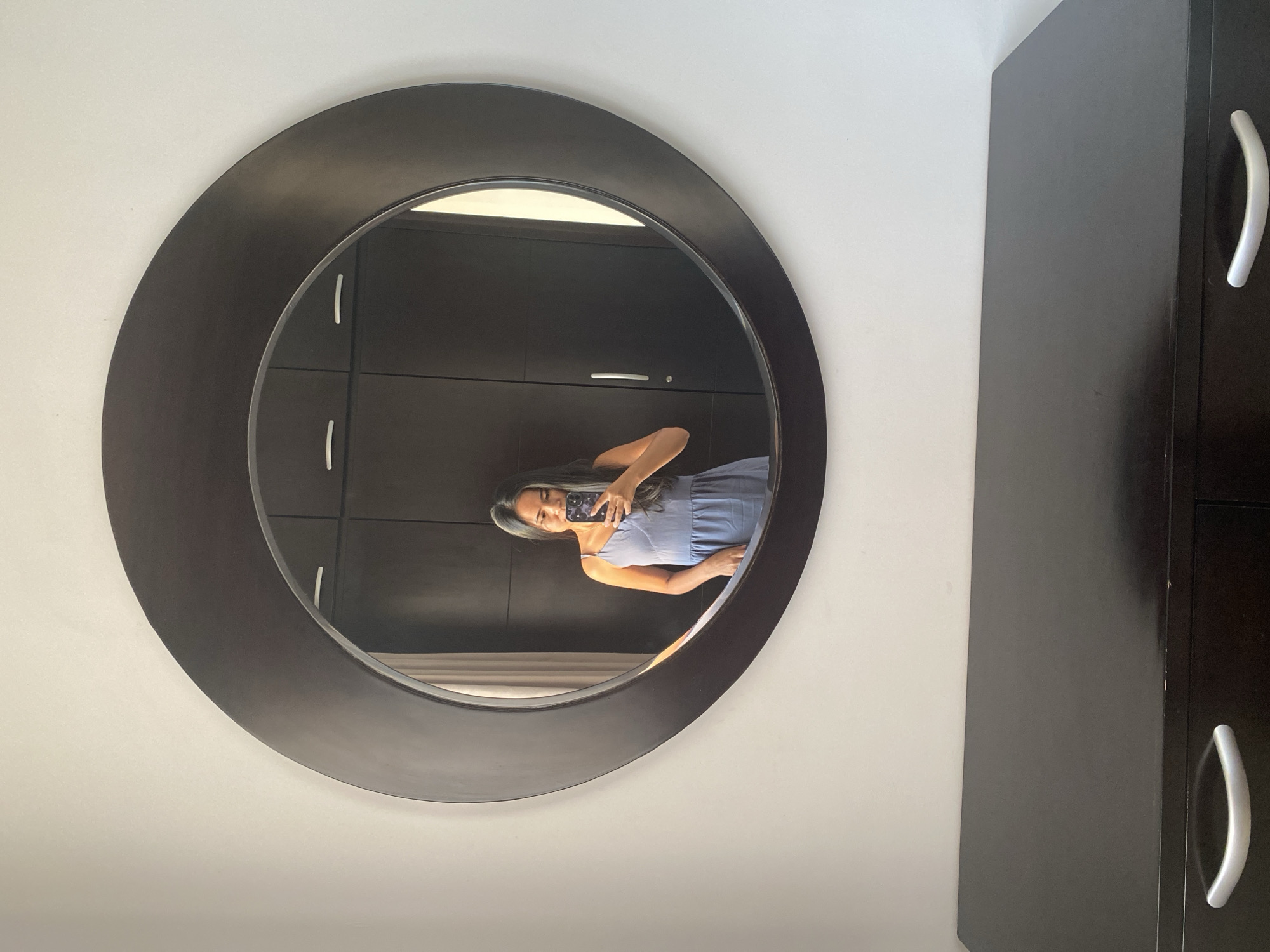 A woman is reflected in a round mirror hanging on a white wall in a room. She is holding a smartphone and taking a selfie. The reflection shows a dark wooden wardrobe with silver handles behind her. The location is likely in North Kuta, Indonesia, based on context clues of the overall image set from which this was derived.