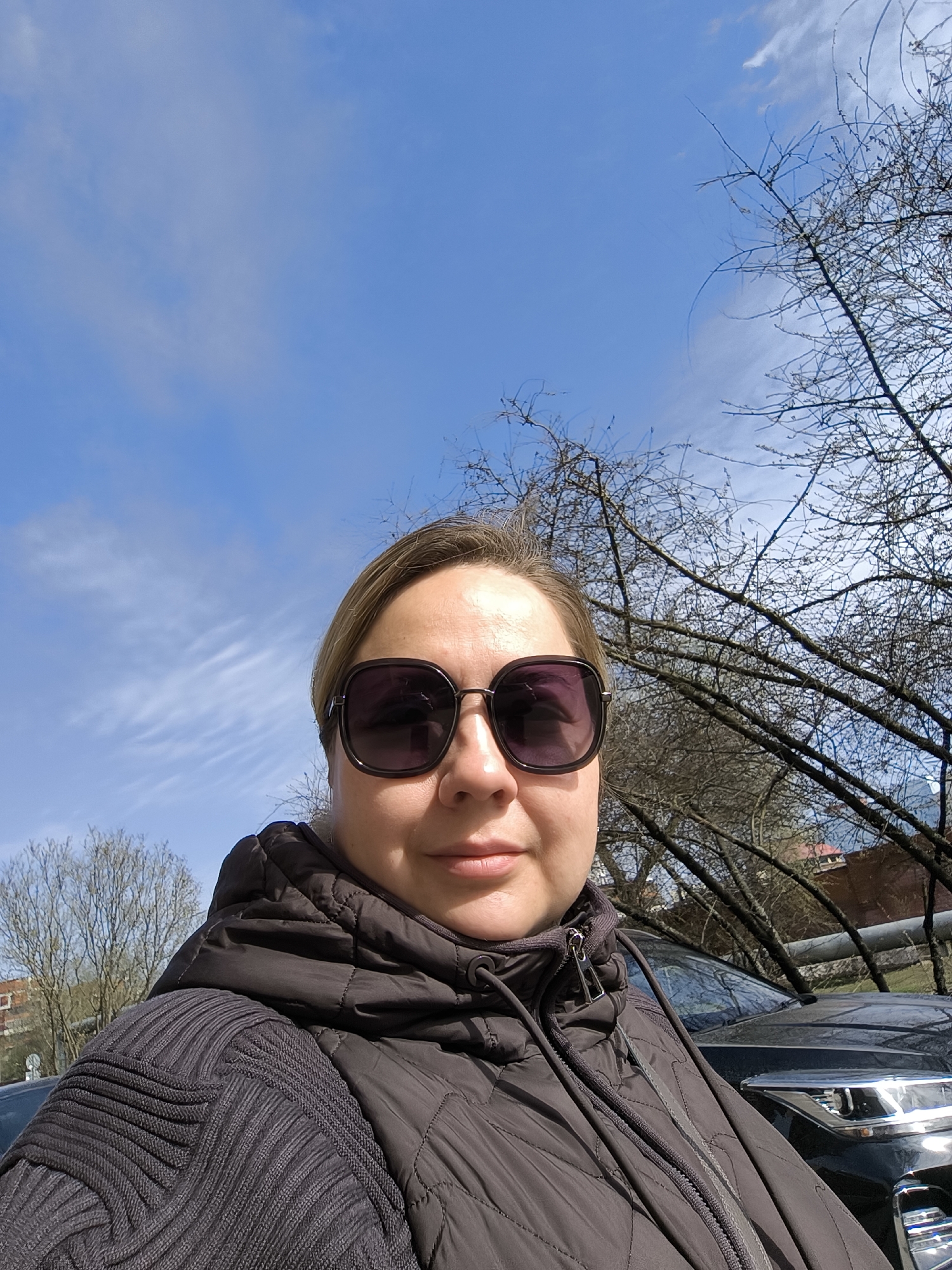 A selfie shows a woman wearing sunglasses and a dark jacket, with a clear blue sky and sparse trees in the background. A building and a black car are partially visible. The scene appears to be outdoors. Based on visual landmarks, the location is likely Khimki, Russia.