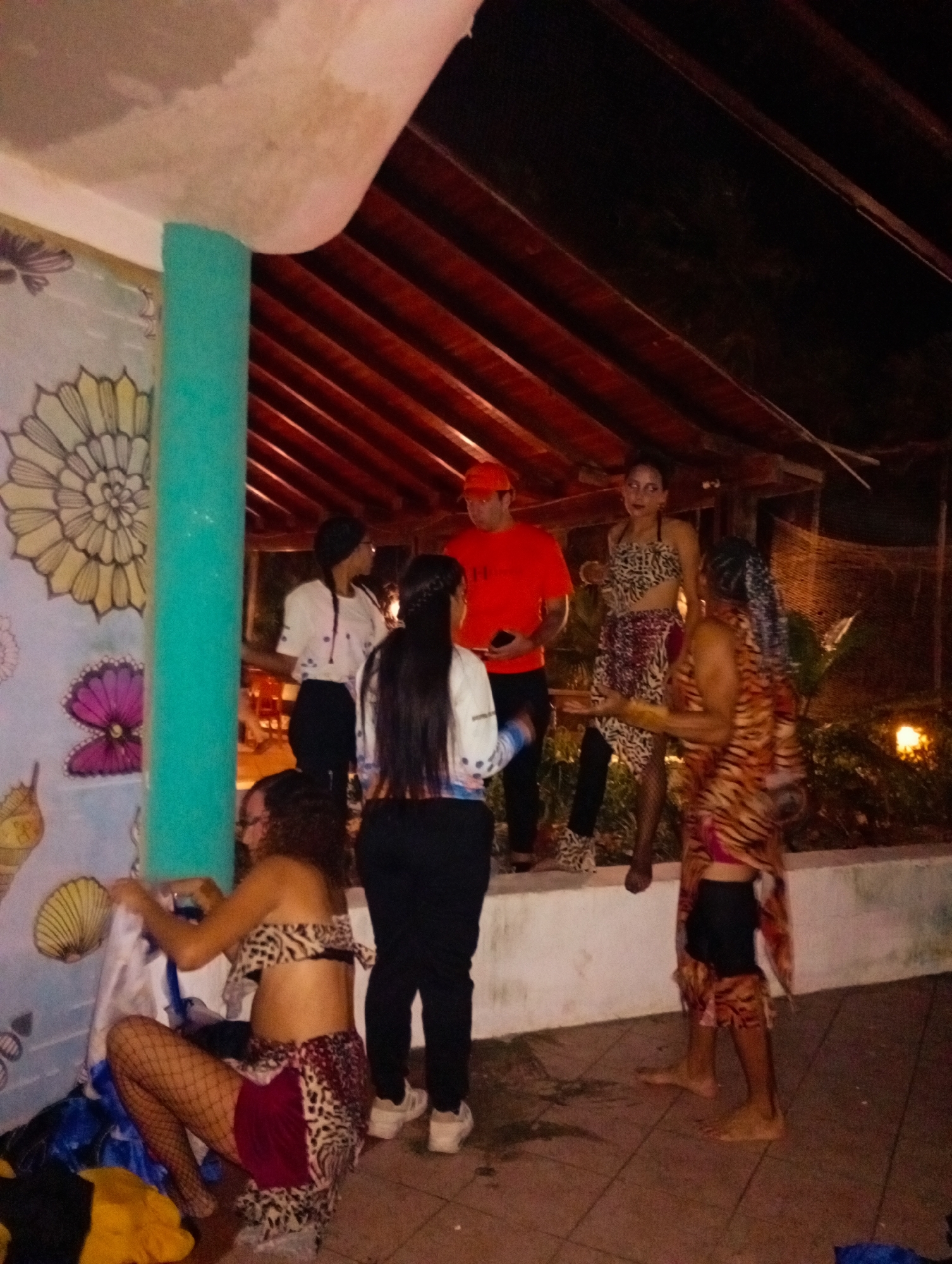 A group of people are gathered at night in what appears to be an outdoor recreational area, possibly near a building with a covered structure. The location appears to be in Playa El Agua, Venezuela. There are seven people visible, some appearing to be wearing costumes or clothing with animal prints. One person is squatting and sorting through items, while another in an orange shirt is standing and facing the group. A person dressed in a tiger-striped outfit stands barefoot. The background shows some foliage and a light. The area is tiled. There is a mural on the wall.