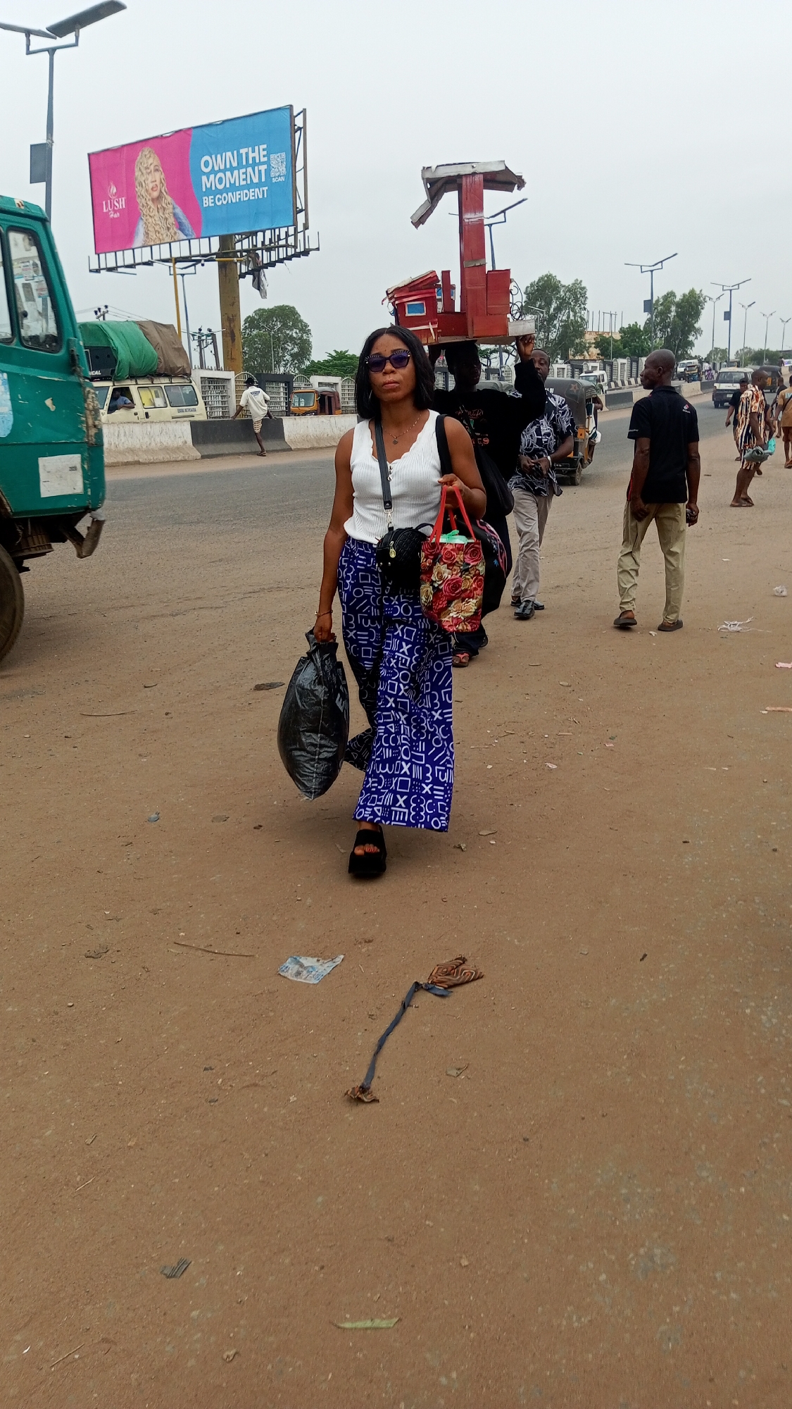 The image captures a bustling street scene in Onitsha, Nigeria, during daytime under an overcast sky. In the foreground, a woman walks towards the viewer on a dusty, unpaved roadside next to an asphalt road. She wears a white sleeveless top, blue wide-leg trousers with a geometric pattern, black platform sandals, and dark sunglasses. She carries a black plastic bag in her left hand, a floral-patterned tote bag in her right hand, and a small black crossbody bag.
In the mid-ground, behind the main subject, several other people are visible. One individual carries a large, red, multi-tiered structure, possibly a mobile vendor's stall, on their head. Further back, pedestrians and various vehicles, including a green truck on the far left and what appears to be a tricycle (keke Napep), line the road. A large billboard stands prominently on the left, displaying an advertisement for "LUSH HAIR" with a picture of a woman with long blonde hair. The text on the billboard reads "OWN THE MOMENT" and "BE CONFIDENT," accompanied by a QR code and the word "SCAN."
The ground beneath the pedestrians is a light brown, unpaved surface with some debris, including discarded paper and a fabric strap. The overall setting suggests a busy urban or semi-urban environment, characterized by pedestrian traffic and commercial activity along a main road.