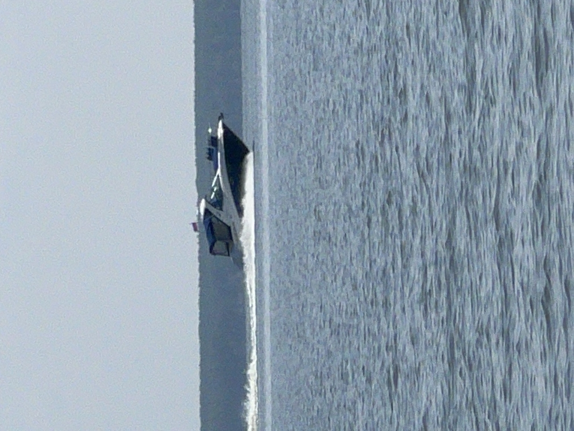 A white motorboat is speeding across the choppy blue water of a lake, leaving a white wake behind it. The boat has a dark blue canopy over the cockpit, and a small Russian flag is flying from the stern. In the background, a dark green, wooded shoreline is visible under a pale, hazy sky. The scene is calm and serene, with no other boats or people visible. This is a photograph of the lake in Borovoe, Russia.