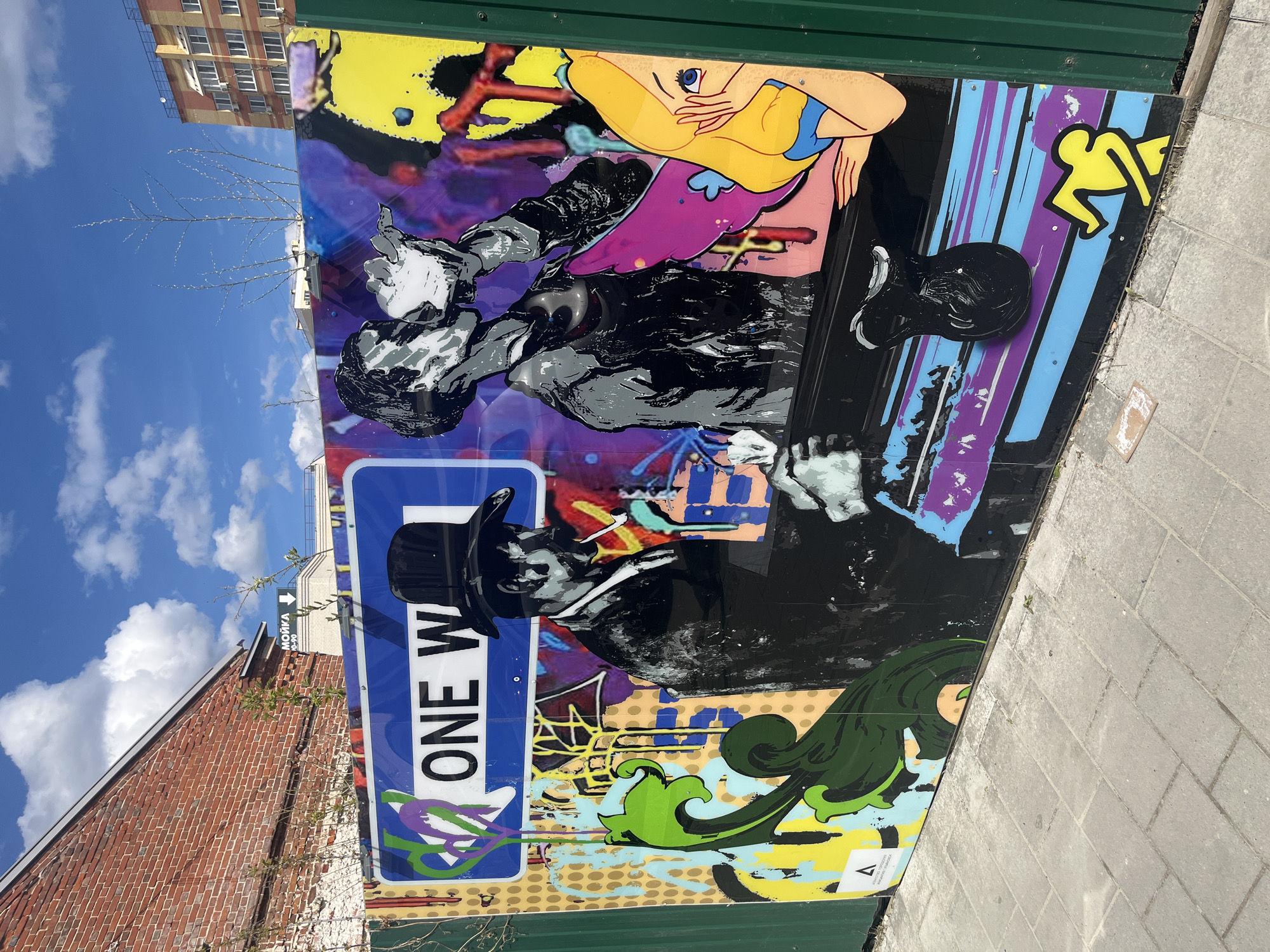 A vibrant street art mural adorns a dark green metal fence or hoarding in an urban setting. The artwork features a blend of styles, including two black-and-white stylized figures reminiscent of classic silent film stars, possibly Charlie Chaplin. One figure, wearing a top hat, is prominently displayed, appearing to smoke a cigar or cigarette. The other figure is partially visible. These are set against a colorful backdrop with abstract splashes of purple, blue, yellow, and orange.
Interspersed with the classic imagery are pop art elements: a blonde cartoon-style woman with large eyes and pink lips, a yellow stick figure, and swirling green decorative patterns. A blue rectangular "ONE WAY" road sign is affixed over the mural, partially overlapping the top-hatted figure.
In the background, under a bright blue sky with scattered white clouds, are several buildings. To the left, a reddish-brown brick building is visible, alongside a bare tree with delicate branches. Further back, a taller building with many windows and an orange-brown facade stands. A white sign with the Russian text "МОЙКА 24/7" (Car Wash 24/7) and an arrow is discernible in the distance. The foreground shows a paved sidewalk or ground. The scene depicts a sunny daytime, highlighting the colorful mural against the urban architecture.