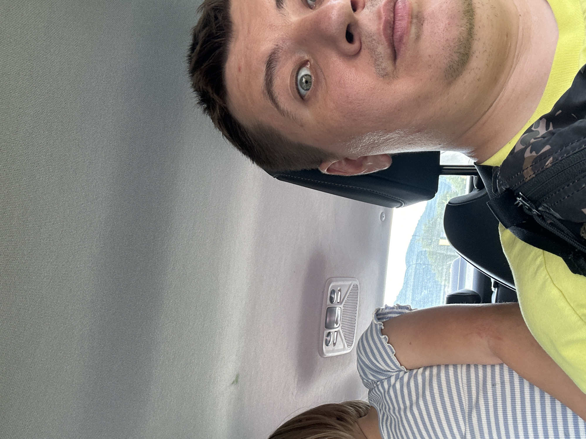 The image shows a close-up view of a man's face as he takes a selfie inside a car. He has light-colored eyes, short brown hair, and stubble on his chin and upper lip. He is wearing a yellow t-shirt and a black bag with a camouflage pattern. In the background, a young person with blonde hair, wearing a blue and white striped shirt, is partially visible. The car's interior, including the ceiling and a light fixture, is also visible. Through the car window, a glimpse of mountains and trees can be seen, suggesting a location outside of a city, possibly in a mountainous region. The image does not provide any specific details to place it in Budva, Montenegro.