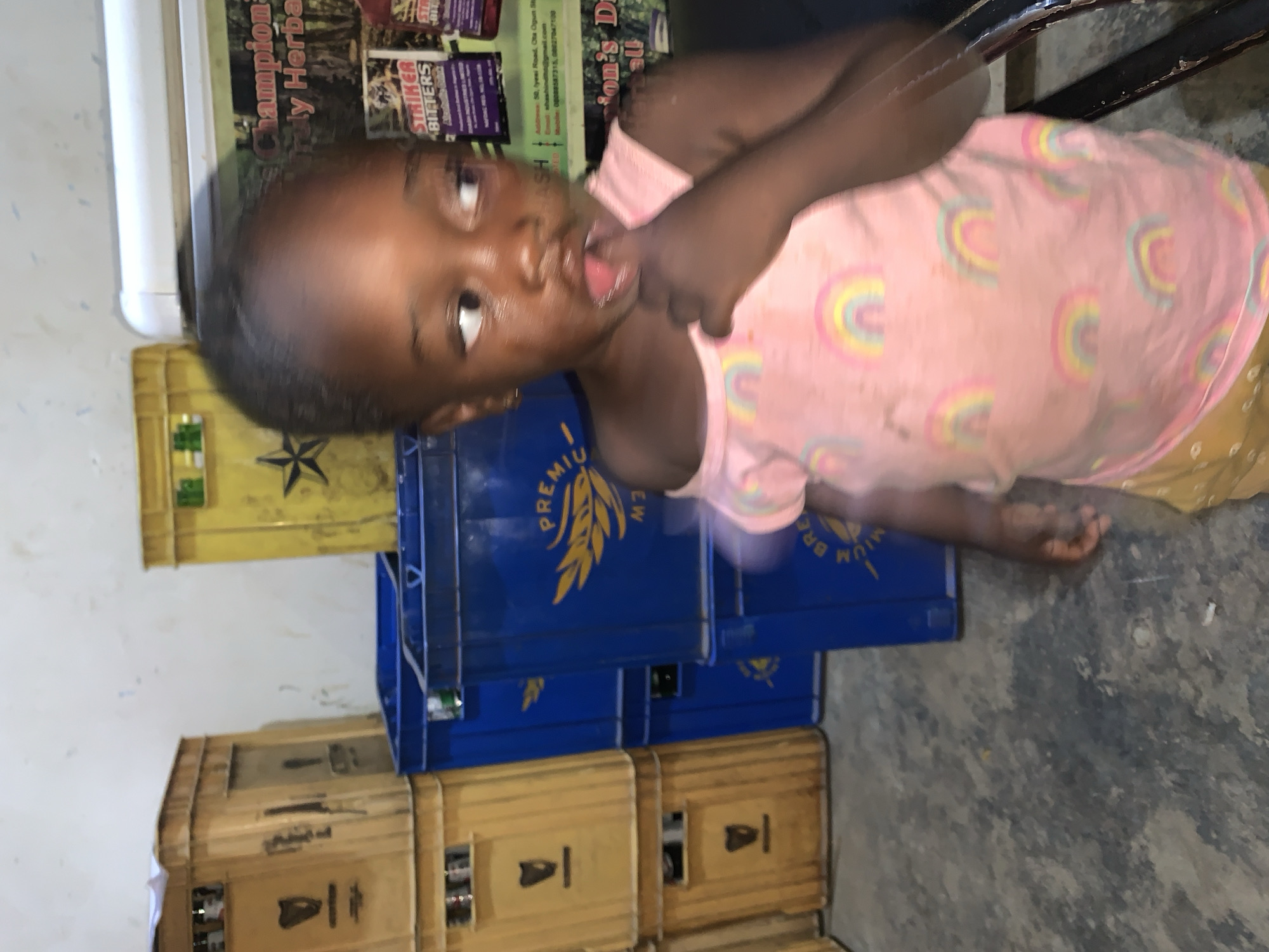 A young child stands in what appears to be an indoor space, possibly a storage area or shop in Kabong, Nigeria. The child, wearing a pink shirt with rainbow patterns, is positioned in front of stacked crates. Some crates are yellow and others are blue, and display branding related to "Premium Brew." The child has a finger in their mouth and is looking toward the camera. In the background is a sign advertising "Champion Herba" and "Striker Bitters," with an address and phone numbers. The floor is gray and appears to be concrete. A portion of what seems to be furniture is visible in the upper right.