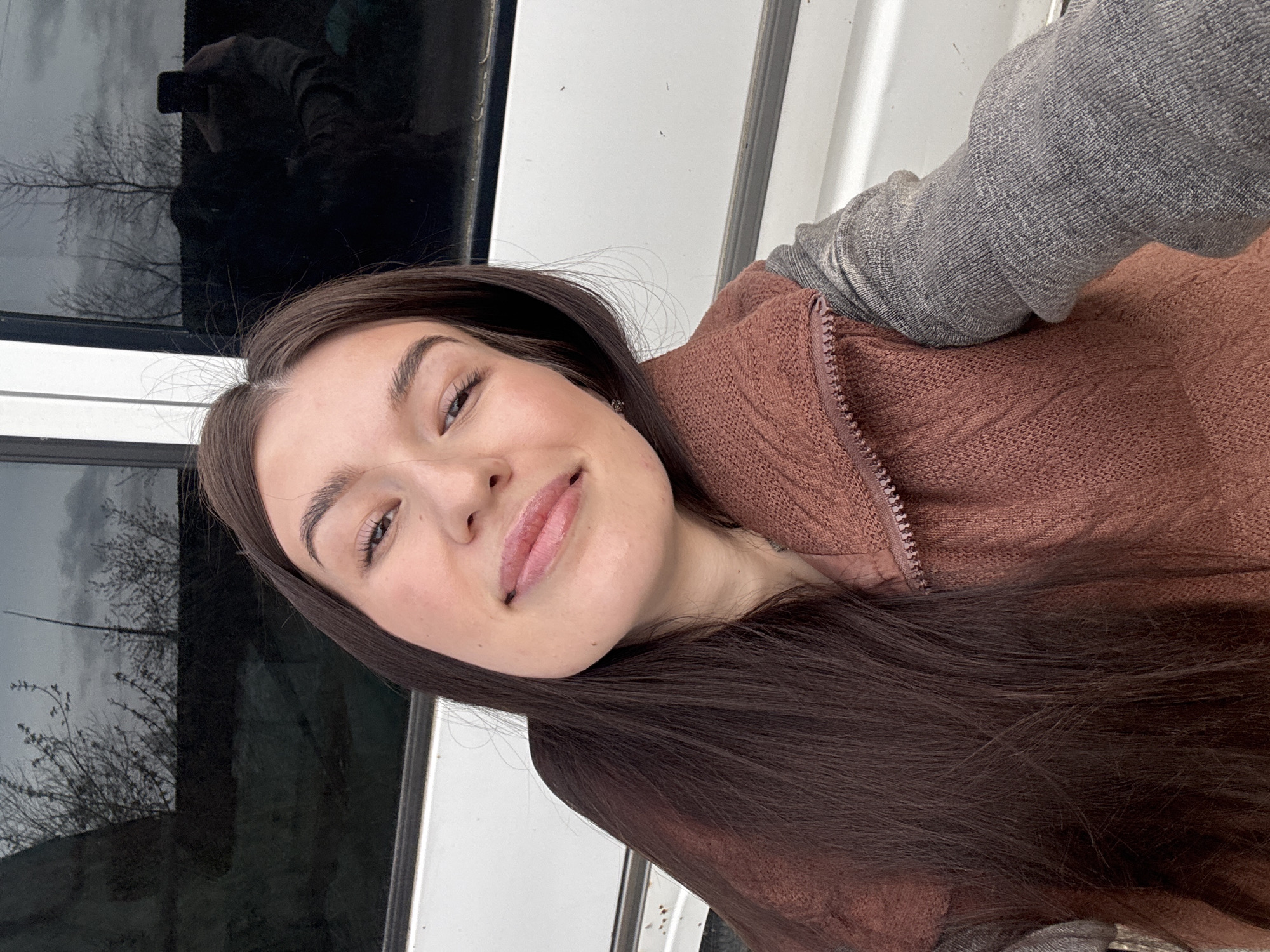 A young woman with long, dark brown hair and fair skin is captured in a close-up portrait, likely a selfie. She has a gentle smile and her head is slightly tilted, looking directly at the viewer. Her dark eyebrows are well-groomed, and she appears to be wearing light makeup, including mascara and a subtle lip gloss. She is dressed in a textured, reddish-brown (russet or mauve) zip-up vest worn over a grey long-sleeved top, suggesting cool weather.
The background is partially obscured by the subject but reveals a dark, reflective surface on her left, which appears to be a window or a glass door panel. Within this reflection, bare tree branches are clearly visible against a light, possibly overcast, sky, indicating late autumn, winter, or early spring. A faint reflection of a person holding a smartphone is also discernible in the glass, suggesting the photograph was taken as a selfie. To her right, a light-colored, possibly white, vertical surface forms part of the background. The lighting is natural daylight, contributing to the soft atmosphere of the image. The scene is set in Chaikovsky, Russia. No visible text is present.