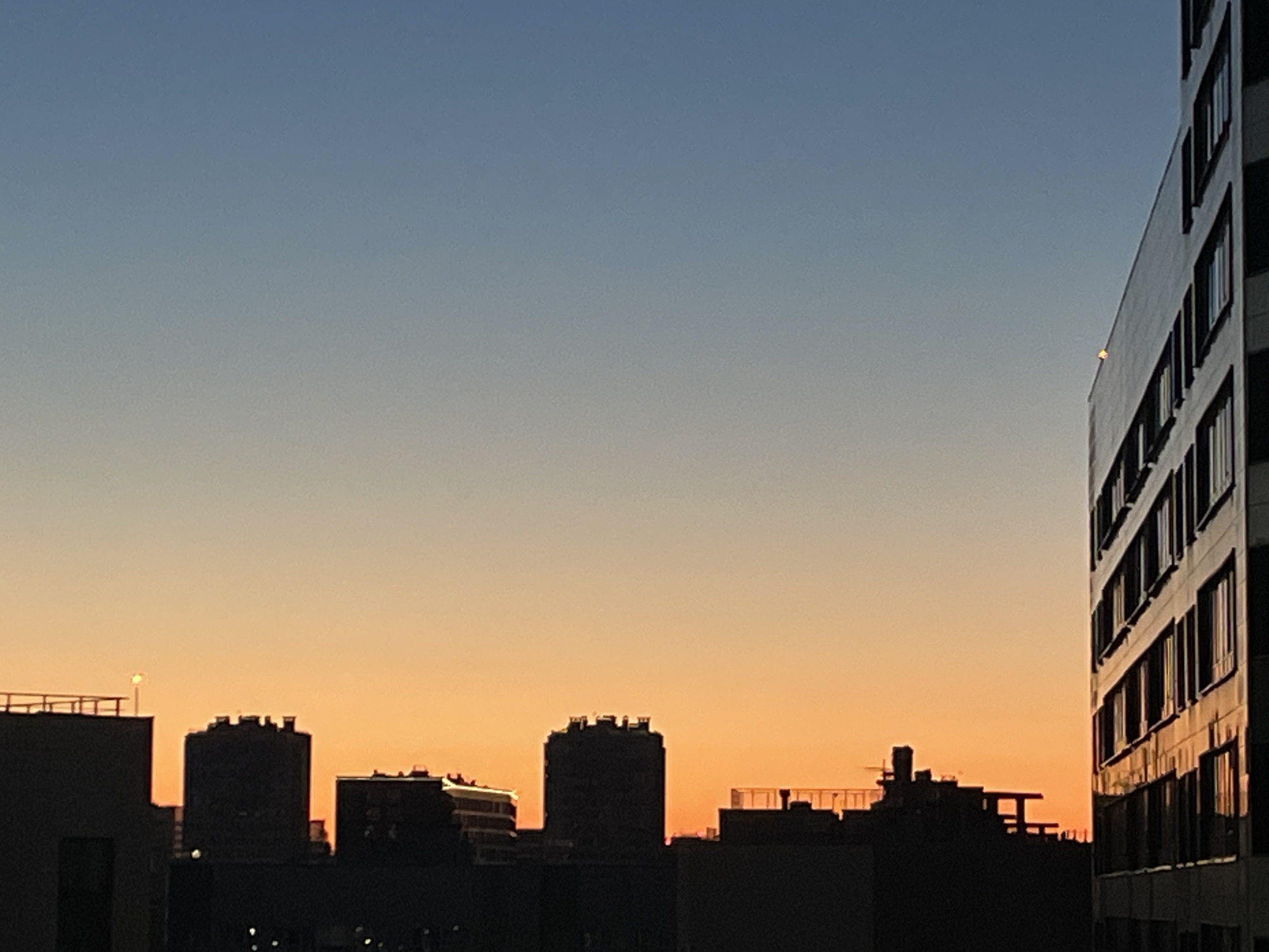 The image captures a city skyline at dusk, with a gradient sky transitioning from a soft blue at the top to a warm orange near the horizon. Silhouetted buildings of various heights and architectural styles are visible against the colorful sky. In the foreground and midground, dark shapes of buildings obscure details, but some rooftop structures and windows can be discerned. A tall, modern building with many windows dominates the right side of the frame, its facade reflecting the evening light. The scene appears to be an urban environment. There are no people or specific actions depicted. The location is identified as Sinyavino, Russia.