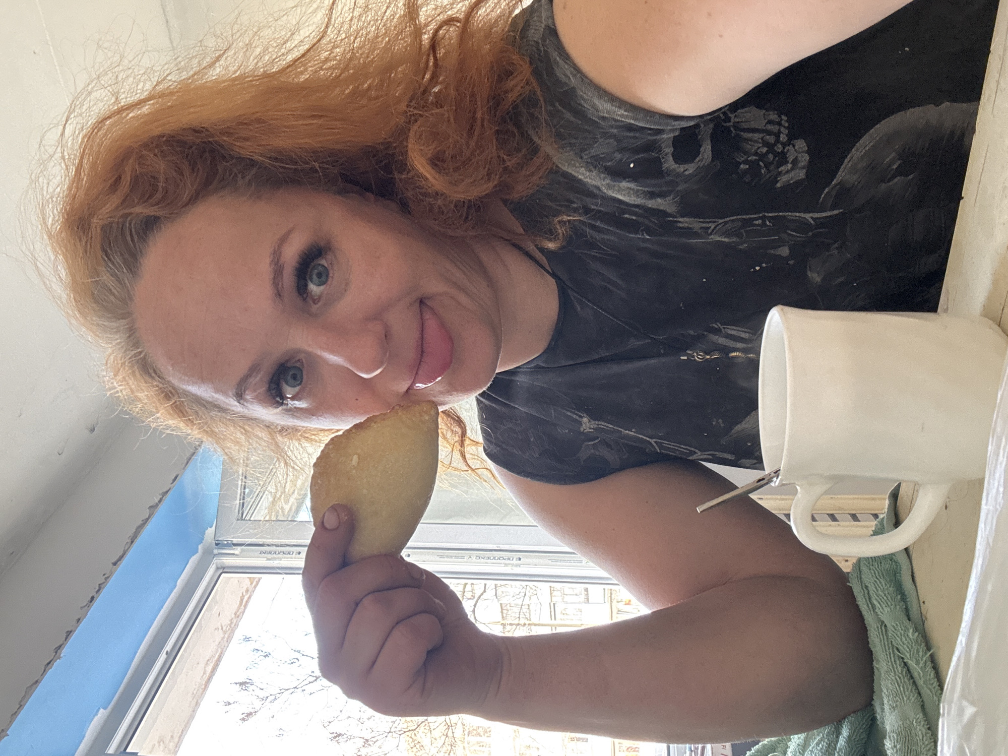 The image presents a woman with long, curly auburn hair and striking blue eyes, captured in a rotated perspective, appearing on her side within the frame. She is looking directly at the viewer with a playful expression, sticking her tongue out slightly towards a small, crescent-shaped pastry held delicately in her left hand. The pastry has a light golden-brown crust, suggesting it is freshly baked.
She is wearing a dark, sleeveless top, possibly grey or black, which features a subtle, lighter-toned pattern that appears abstract or graphic, with hints of skeletal or bone-like forms near the shoulder.
To her right, a white ceramic mug with a visible handle holds a metallic spoon. A light green towel or cloth is draped beneath or beside the mug. The background reveals a window, through which natural daylight streams in, illuminating bare tree branches outside. The white frame of the window displays partially visible text that reads "DECODENS" and another segment that appears to be "PPCNNEC". Adjacent to the window, a section of a wall or surface painted in a distinct blue color is visible, with some rough edges suggesting it might be under renovation or decoration. The scene depicts a casual, lighthearted moment, likely a snack break during daytime in an indoor setting in Chaikovsky, Russia.