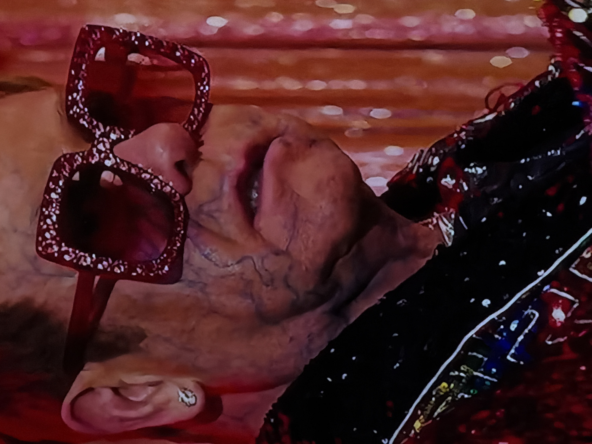 The media file presents a close-up, angled view of a person, likely an adult, in a dramatic setting. The visible subject includes the right side of their face, featuring a cheek, part of the nose, and an ear adorned with a small, circular earring. The skin shows visible veins, particularly on the side of the face, suggesting either an older age or specific lighting conditions.
The person is wearing large, square-framed sunglasses that are a prominent feature. The frames are deep red and heavily embellished with numerous small, sparkling elements, indicating an ornate and possibly theatrical design. These sunglasses rest high on the nose, partially obscuring the eyes.
A portion of what appears to be an elaborate, dark garment or costume with red accents is visible on the right side of the frame. This attire, combined with the sparkling sunglasses, suggests a performance, a festive event, or a highly stylized photo shoot. The individual appears to be resting or posing, as indicated by their reclined posture.
The background is softly blurred, creating a bokeh effect with warm, reddish-orange hues and indistinct points of light. This lighting and atmospheric blur strongly imply an indoor setting or an evening event, such as a stage, a club, or a party environment. No specific activity beyond posing or resting is discernible.
There is no visible text in the image. The overall impression is one of flamboyant style and a moment of quiet repose within a vibrant, possibly glamorous, context. This scene is situated in Sofiivska Borshchahivka, Ukraine.