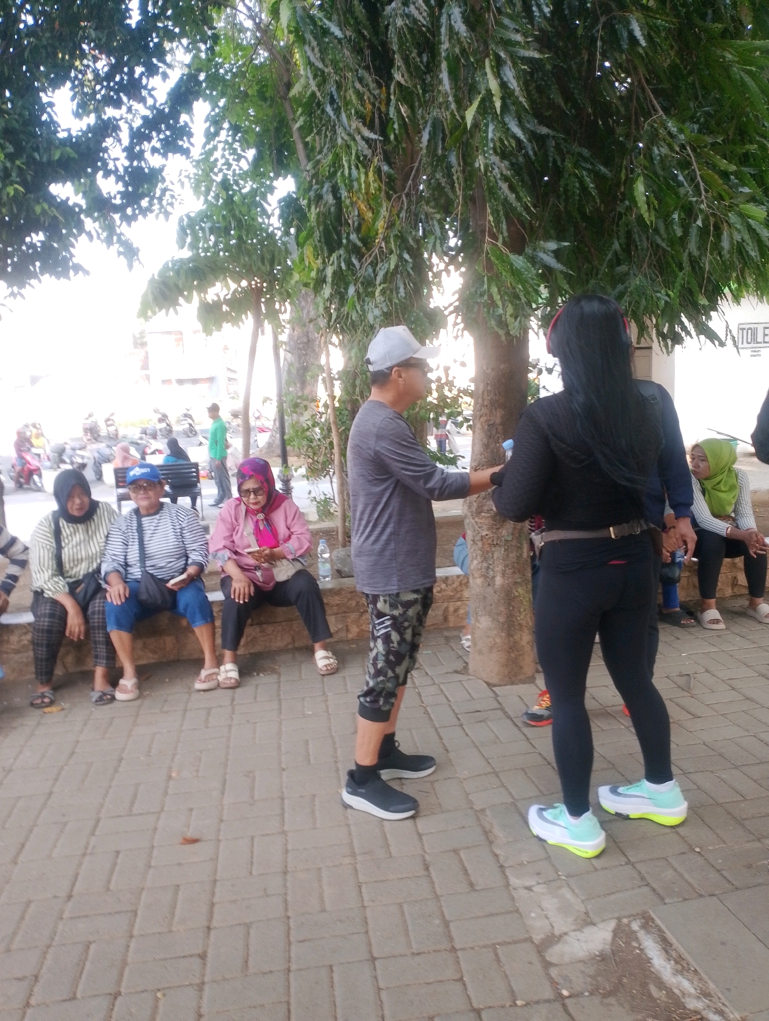 The image depicts an outdoor scene likely in Probolinggo, Indonesia, with several people gathered near trees and a paved walkway. In the foreground, a man in a gray t-shirt and camouflage shorts stands, facing away from the camera, talking to a person with long black hair wearing a black outfit and red headphones. To the left, three women are seated on a low concrete wall. The woman on the far left wears a striped shirt and patterned pants. The middle woman is in a striped shirt and blue pants, wearing a blue baseball cap. The woman on the right is dressed in a pink top and black pants, holding a smartphone. Behind them, motorcycles are parked, and other individuals can be seen in the background, including a person in a green shirt standing further away. A "TOILET" sign is visible on a white building in the upper right corner. The environment appears to be a public area with trees, a paved walkway, and seating.