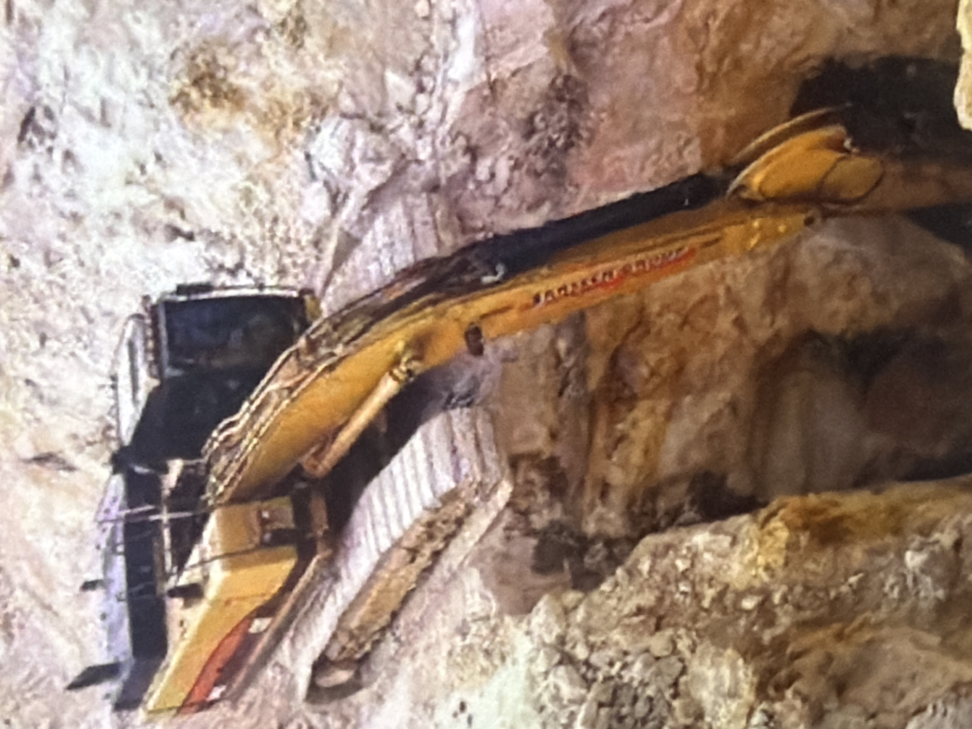 An excavator is positioned in a trench, with its tracks partially submerged in dirt. The excavator is yellow and red, with a long arm reaching down into the trench. The surrounding environment is rocky and consists of light brown and beige soil. The scene is likely a construction or mining site. The location is identified as Bukuru, Nigeria. There are no people visible in the image.