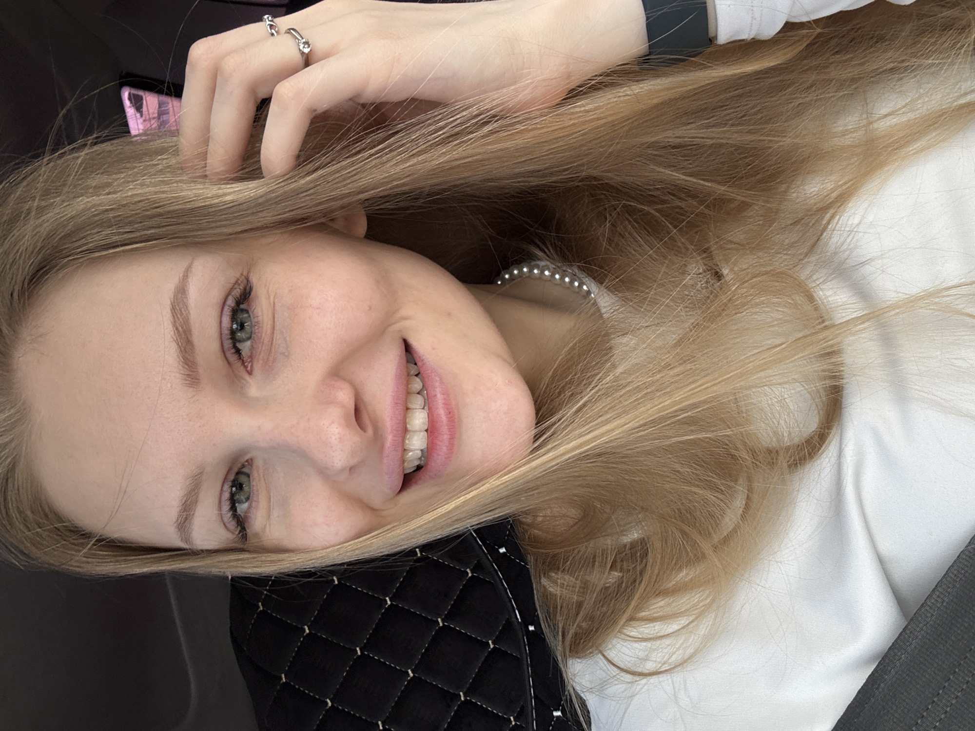A close-up, rotated selfie captures a young woman, likely in her late teens or early twenties, inside what appears to be a vehicle in Perm, Russia. Her long, light blonde or light brown hair flows around her face and shoulders. She has fair skin, prominent green eyes, and a radiant smile that reveals her teeth, with visible braces or aligners on the lower set.
Her right hand, adorned with two silver rings on her index and middle fingers, is casually positioned in her hair. She is wearing a white top, and a pearl necklace is partially visible around her neck. A watch or fitness tracker is noticeable on her left wrist.
The setting is an interior space, strongly suggested to be a car by the presence of a dark gray seatbelt strap in the bottom right corner and dark background elements. A black quilted object, possibly a bag or cushion, rests near her head, and a hint of a pink object can be seen behind her on the left. The lighting is soft and even. The woman's expression conveys happiness, cheerfulness, and a relaxed demeanor. No visible text is present.