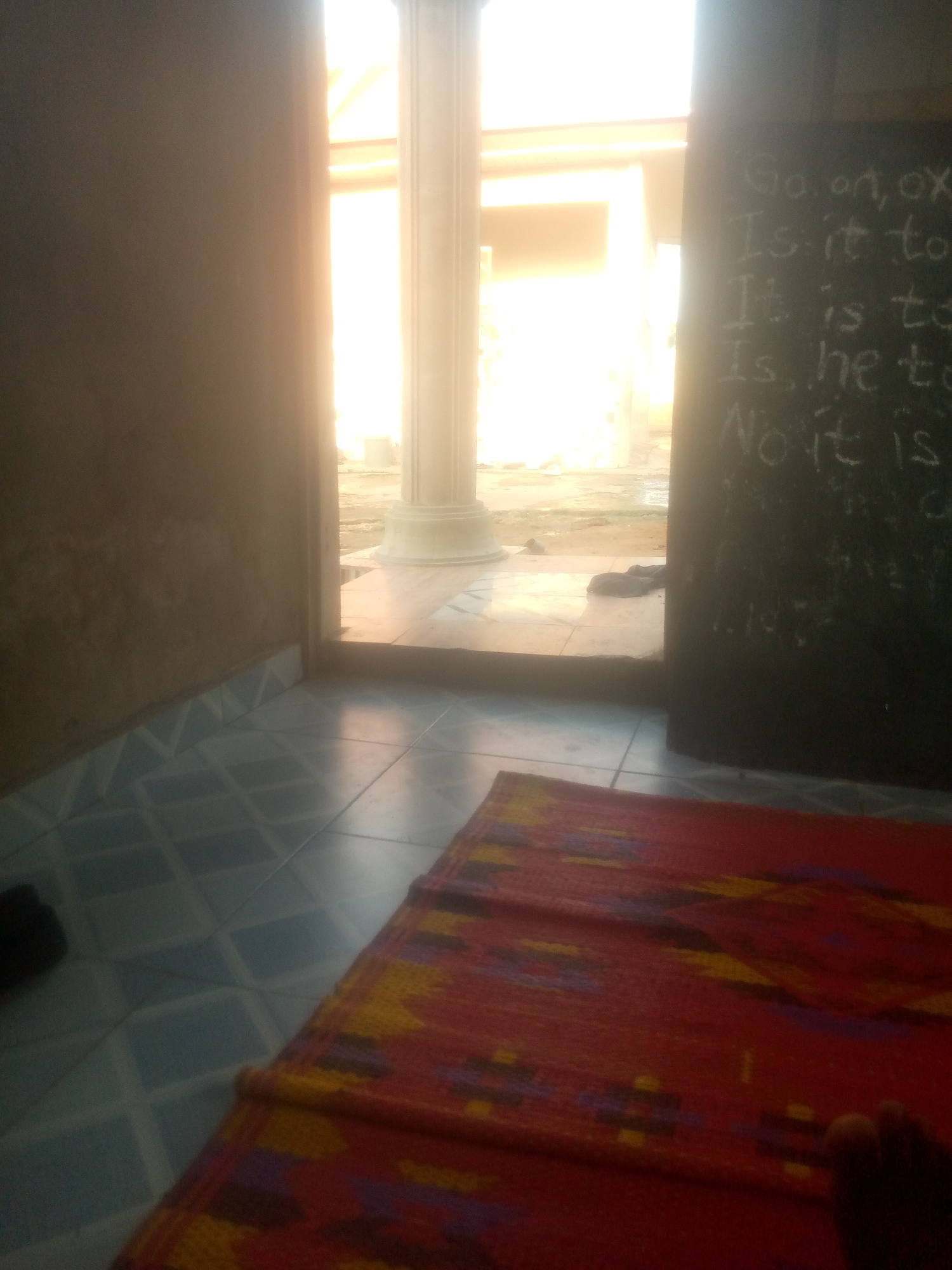 The image depicts an indoor scene, likely a classroom or study area, in Jauro Yinu, Nigeria. The floor is tiled with a blue diamond pattern. A red and yellow patterned mat lies diagonally across the foreground. To the right, a dark surface, possibly a chalkboard or whiteboard, displays white chalk writing. The writing appears to be a series of sentences, some starting with "Is it to" and one with "No it is". Sunlight streams in from an open doorway or window in the background, illuminating a large pillar and the outdoor environment. The overall lighting is bright and somewhat overexposed, particularly in the background.