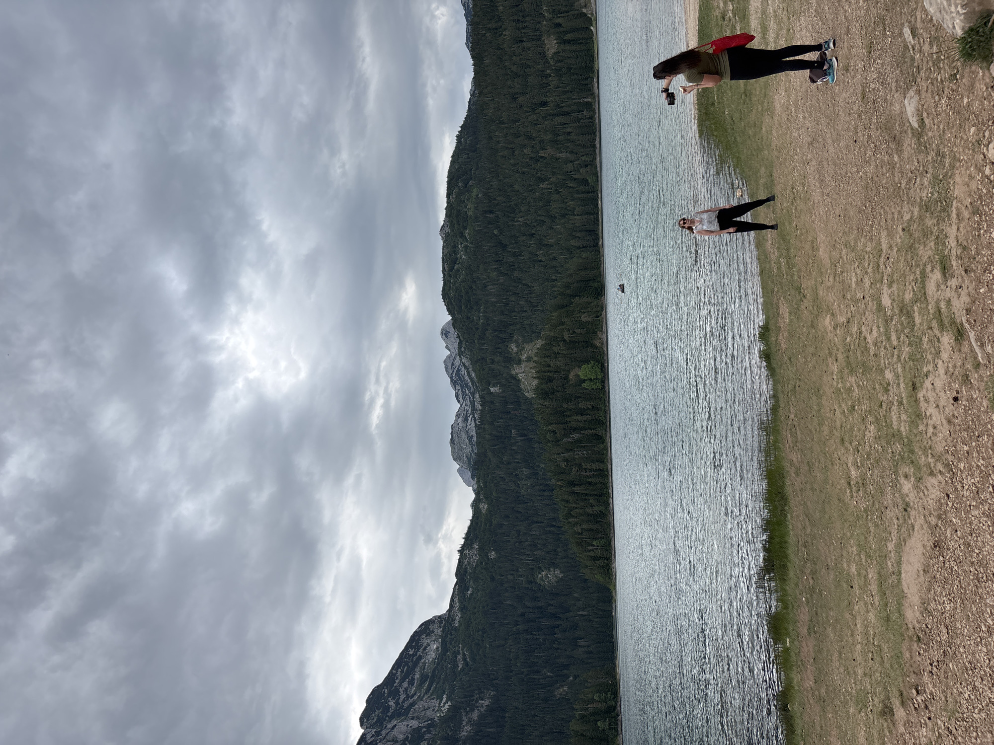 The image captures a serene natural landscape in Pitomine, Montenegro, featuring a vast lake bordered by a dense, dark green forest that climbs up rugged, grey mountains under a cloudy sky. In the foreground, a dry, sandy area with scattered pebbles transitions to a grassy bank along the lake's edge. Two women are present on the shore. One woman, on the right, is holding a video camera, appearing to film the other woman who is standing on the grass to her left, facing the camera. A small boat is visible on the water in the distance. The overall atmosphere is calm and picturesque, with the overcast sky lending a soft, diffused light to the scene.