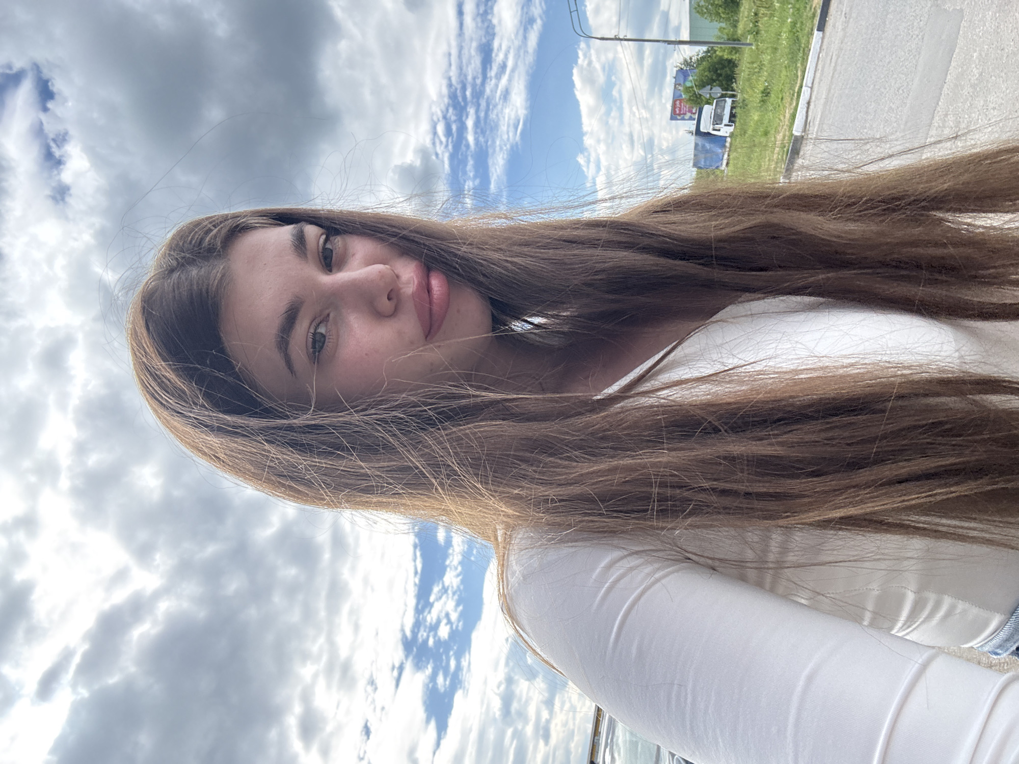 The image is a portrait of a young woman taken outdoors. She is positioned in the foreground, her face tilted slightly upwards and to the left. Her long, brown hair is a prominent feature, cascading down around her and partially obscuring the background. She is wearing a white, long-sleeved top.
The background consists of a cloudy sky with patches of blue. To the right, a paved road is visible, with a grassy verge and some trees and buildings in the distance. A white van and a billboard are also discernible in the background, suggesting an outdoor, possibly rural or roadside, location. The lighting suggests it is daytime. The woman's expression appears neutral or relaxed. There is no visible text in the image. The metadata indicates the location is Troshkovo, Russia.