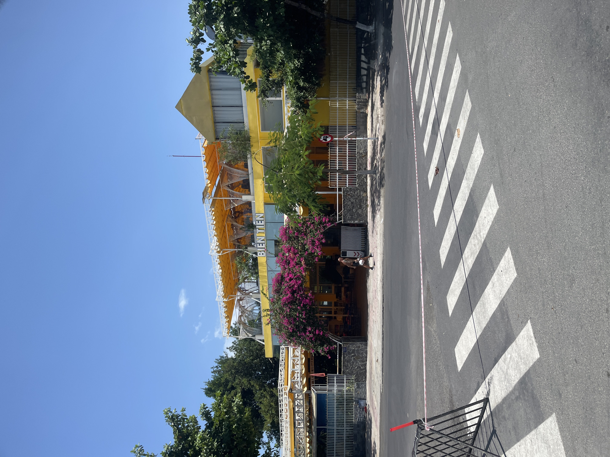 The image shows a bright yellow building in Nha Trang, Vietnam, with a prominent "BIỆN TIÊN" sign. The building has multiple levels, with awnings on the upper floors and an abundance of pink bougainvillea flowers cascading down the facade. A pedestrian crosses the street in front of the building on a clearly marked crosswalk. The street is paved, and there are trees lining the sidewalk. A temporary fence with red and white tape is visible near the crosswalk. The sky is clear and blue, indicating a sunny day.