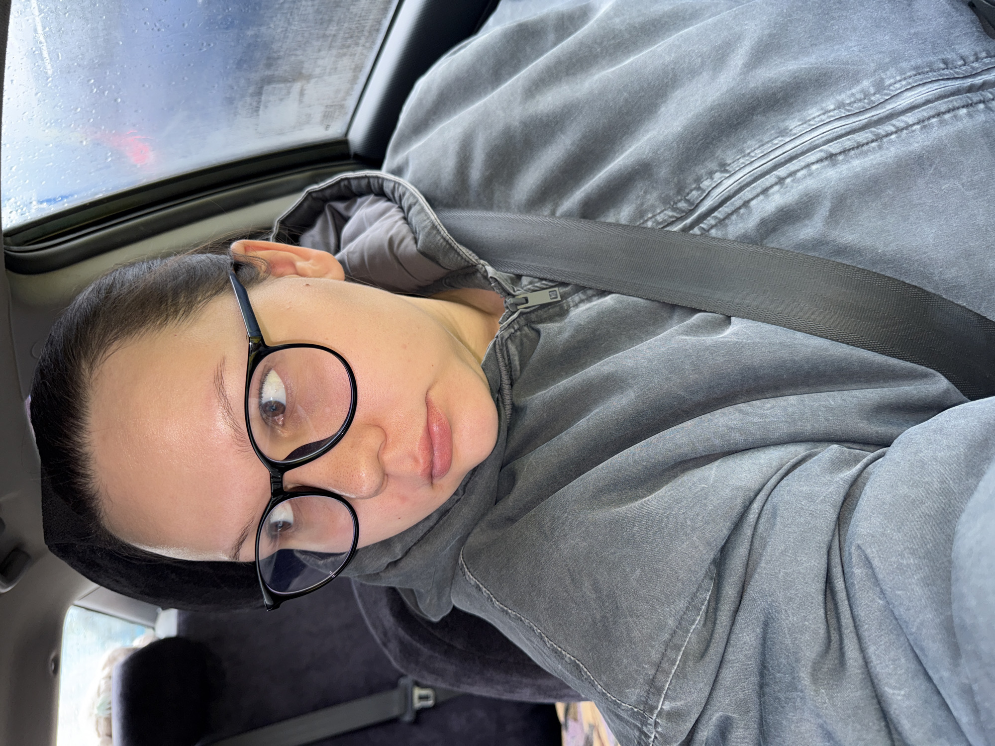 A person is in a car, wearing a gray hooded jacket and black glasses, with a seatbelt fastened. The scene appears to be inside a car, with a sunroof visible and rain drops on the glass. The car is located in the city of Chaikovsky, Russia.