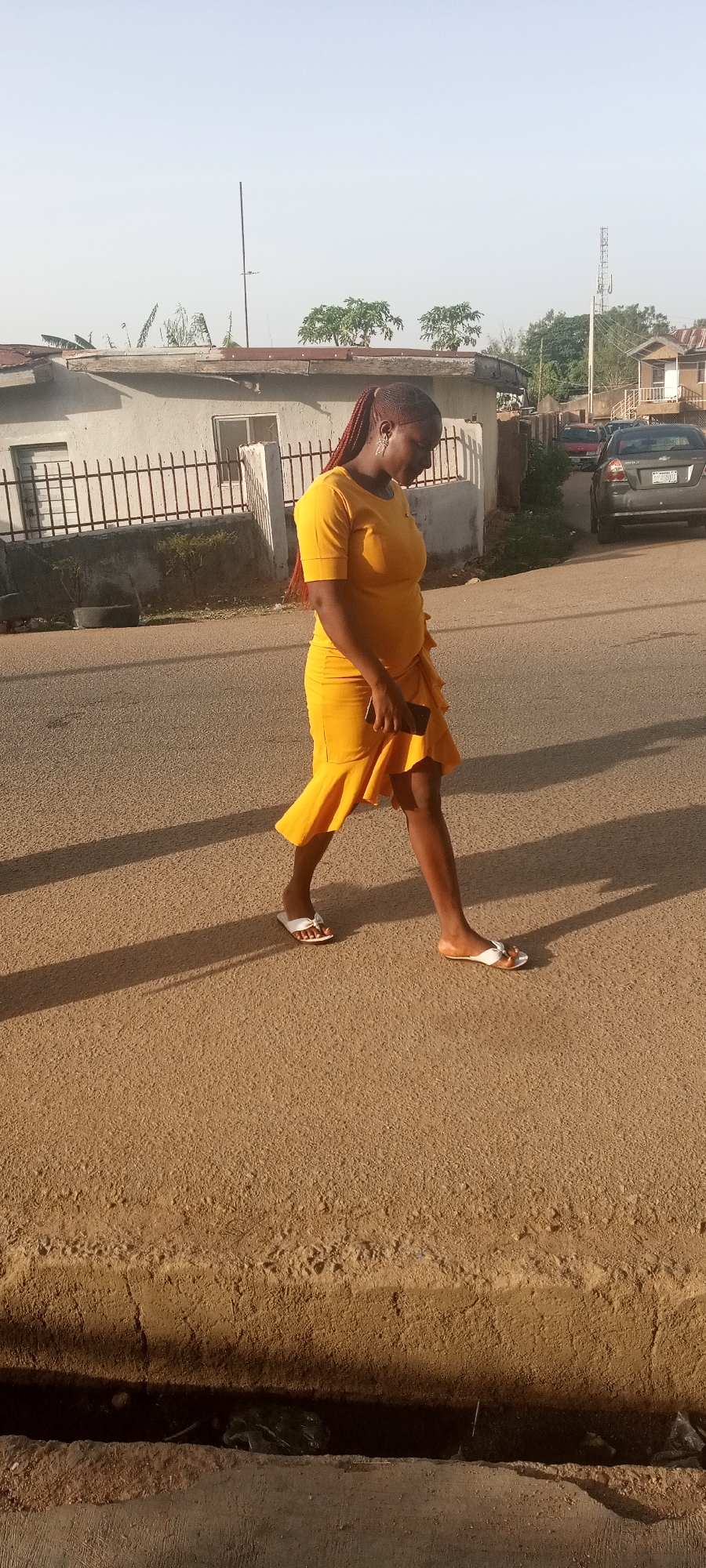 A woman with dark skin and braided hair walks on an asphalt road. She is wearing a yellow dress and white sandals. She is carrying a black phone in her hand. There are houses and parked cars in the background. The location is likely a residential area in Jos, Nigeria, based on the architecture and environment. The time of day appears to be early morning or late afternoon due to the long shadows cast on the road.