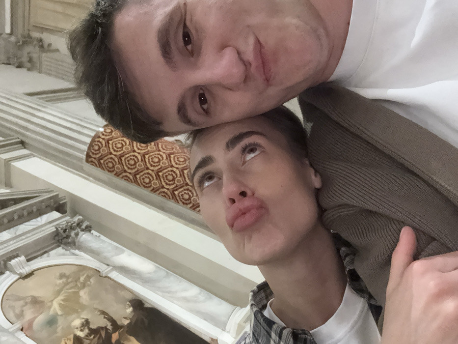 A selfie captures two individuals in what appears to be the interior of the **Basilica dei Santi Giovanni e Paolo (San Zanipolo)** in Venice, Italy. The image is tilted. On the left, a female individual with dark hair and pronounced eyebrows is depicted looking upwards with pursed lips, forming a "duck face" expression. She wears a white t-shirt layered under a plaid shirt and an olive-brown ribbed sweater, and rests her head on the shoulder or chest of the male individual. On the right, a male individual with short dark hair faces the camera, also displaying pursed lips in a similar pouting expression. He is wearing a white t-shirt. The background features grand architectural elements: a white marble column with an ornate capital is visible to the upper left. Above and behind the female, a portion of a ceiling or wall fresco depicting classical figures in a celestial scene is discernible. Adjacent to the column and behind both individuals is a section of wall or pillar covered with a reddish-brown patterned fabric featuring light, circular geometric motifs. The overall environment suggests a richly decorated, historic indoor space, consistent with a major Venetian church.