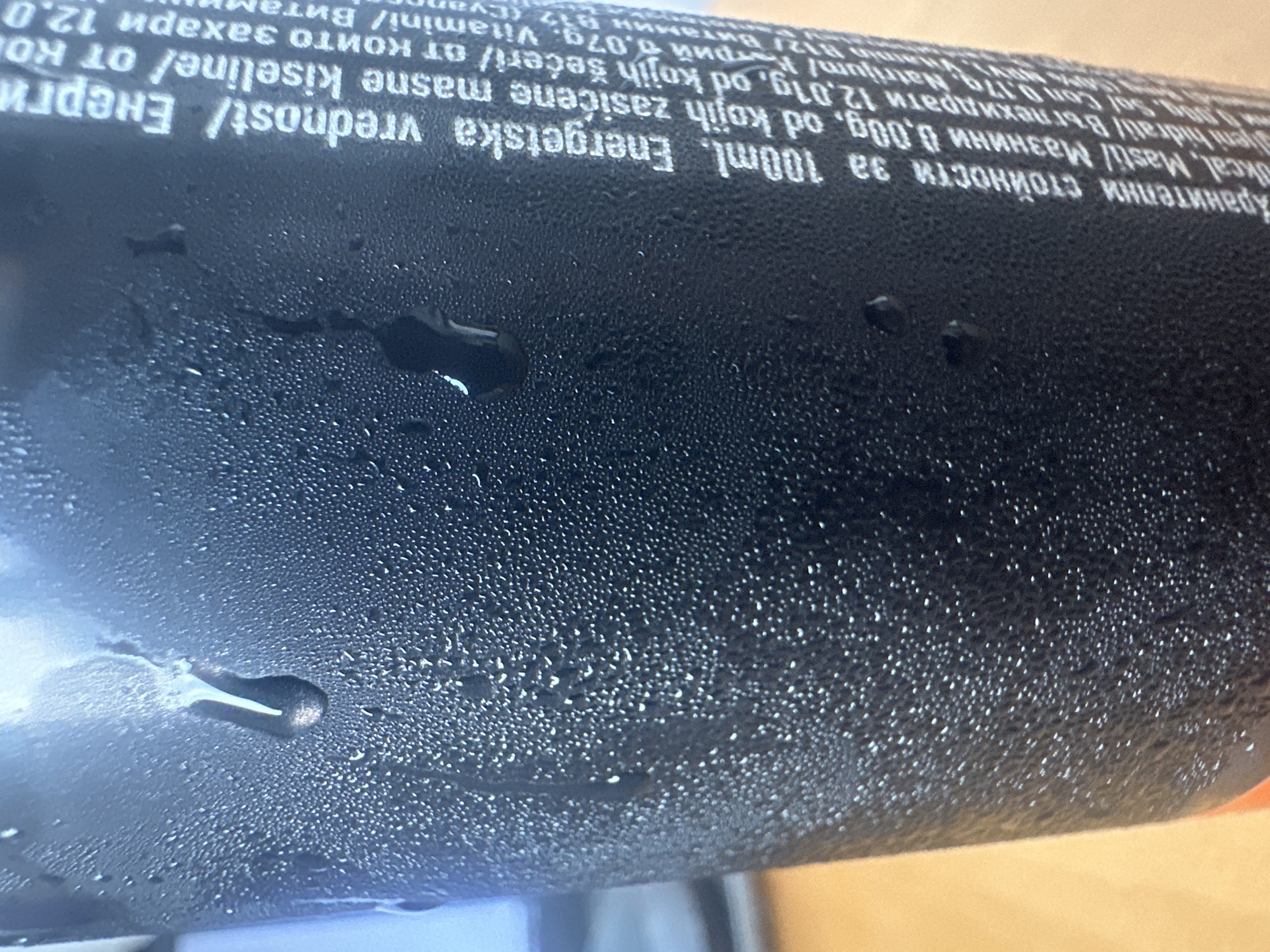 The image shows a close-up of a black can covered in condensation. The can has text printed on it, which appears to be nutritional information. The text is white and oriented vertically, running from bottom to top. Part of the text is in a language that appears to be Cyrillic, with some Latin characters mixed in. The environment appears to be indoors, with a blurred background that suggests a light-colored surface and possibly a screen or monitor. There are no people visible in the image. The image is a still photograph, capturing a moment in time. The location is not identifiable from the image itself.