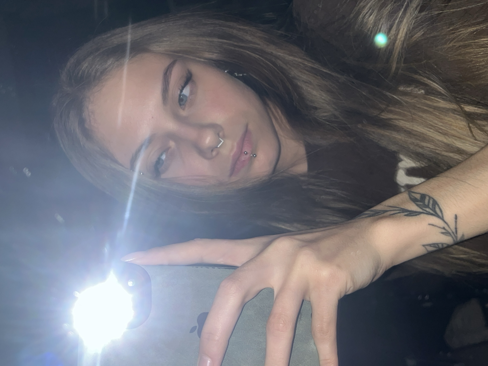 A young woman is taking a selfie in a low-light environment using the flash of her smartphone. She has light brown hair, fair skin, and facial piercings, including a septum ring and a lip piercing. A tattoo of a leafy branch is visible on her wrist. The phone's flash creates a bright glare, partially obscuring the phone itself. The location is indiscernible.