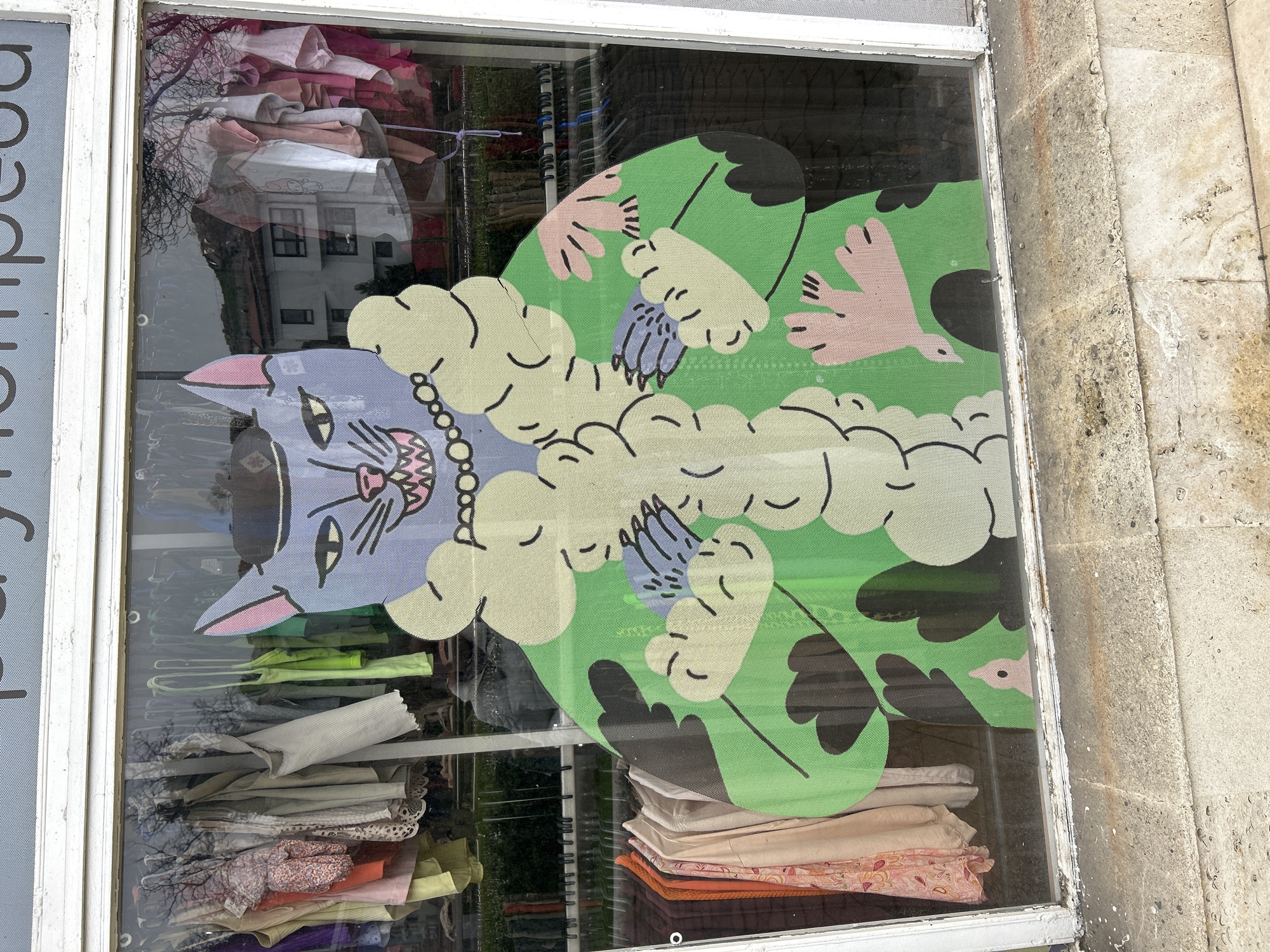 A slightly tilted, eye-level photograph shows a window display in Plovdiv, Bulgaria. The window is framed in white-painted wood. A large, graphic, stylized cat rug is partially visible in the window. The cat's head is light purple, with pink details and wearing a string of pearls. The cat lies on a green background with black and pink accents, appearing to be a landscape. Fluffy cream-colored cloud shapes are also part of the design. Beneath the rug, stacks of neatly folded clothing in various colors are visible. Garments on hangers are visible higher up in the window. The window reflects a light-colored building with windows and a tree. A partial sign with Cyrillic letters is on the left side of the window frame. The outside wall to the right of the window is made of light brown stone blocks.